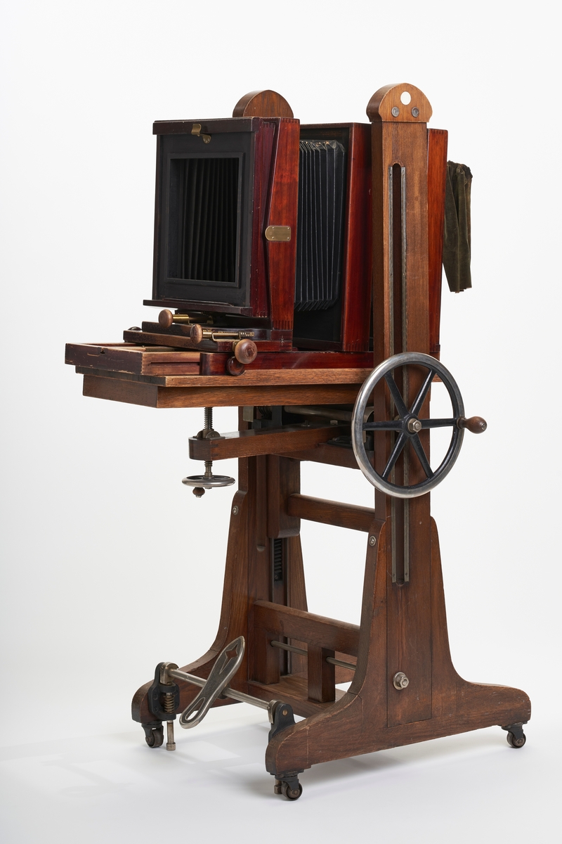 Fotografiapparat på stativ. Stativet er massivt og har justerbar sokkel. Både fotografiapparatet og stativet er i tre. Stativet står på hjul. Fotografiapparatet har uttrekkbar belg, men mangler frontplate og opptikk. Det hører med en slange til fotografiapparatet og en innstikksmodul for å velge blenderåpnimg på motivet.