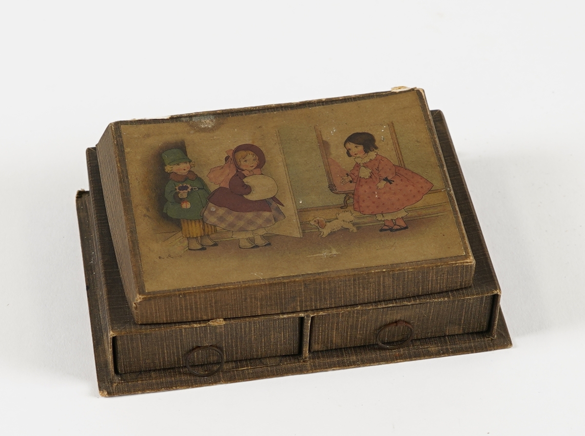 Ei rektangulær øskje med tre rom; to skuffer nedst og ei flat øskje på toppen. I skuffene er det små papirkonvoluttar med raudt band rundt, og i toppen er det små, linjerte ark. I øskja på toppen er det òg diverse små "skattar"; eit teikna portrett, eit utklypt bilete av ein påfugl, eit lite perlearmband (+to lause perler), nokre små korallbitar og ein kalendarlapp (Oktober 12 Søndag). På loket på toppen er det bilete av tre dokkeliknande figurar, og ein liten hund.