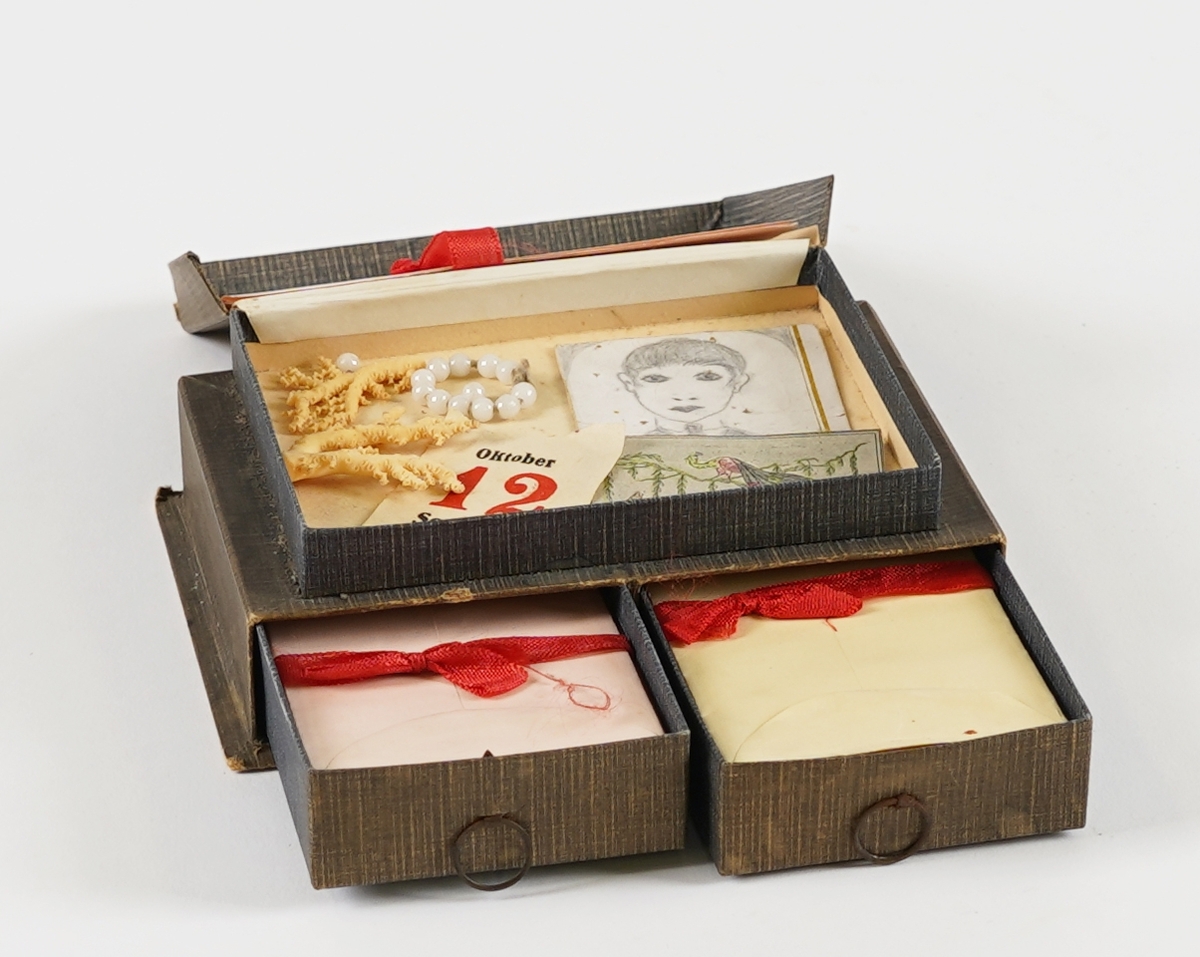 Ei rektangulær øskje med tre rom; to skuffer nedst og ei flat øskje på toppen. I skuffene er det små papirkonvoluttar med raudt band rundt, og i toppen er det små, linjerte ark. I øskja på toppen er det òg diverse små "skattar"; eit teikna portrett, eit utklypt bilete av ein påfugl, eit lite perlearmband (+to lause perler), nokre små korallbitar og ein kalendarlapp (Oktober 12 Søndag). På loket på toppen er det bilete av tre dokkeliknande figurar, og ein liten hund.