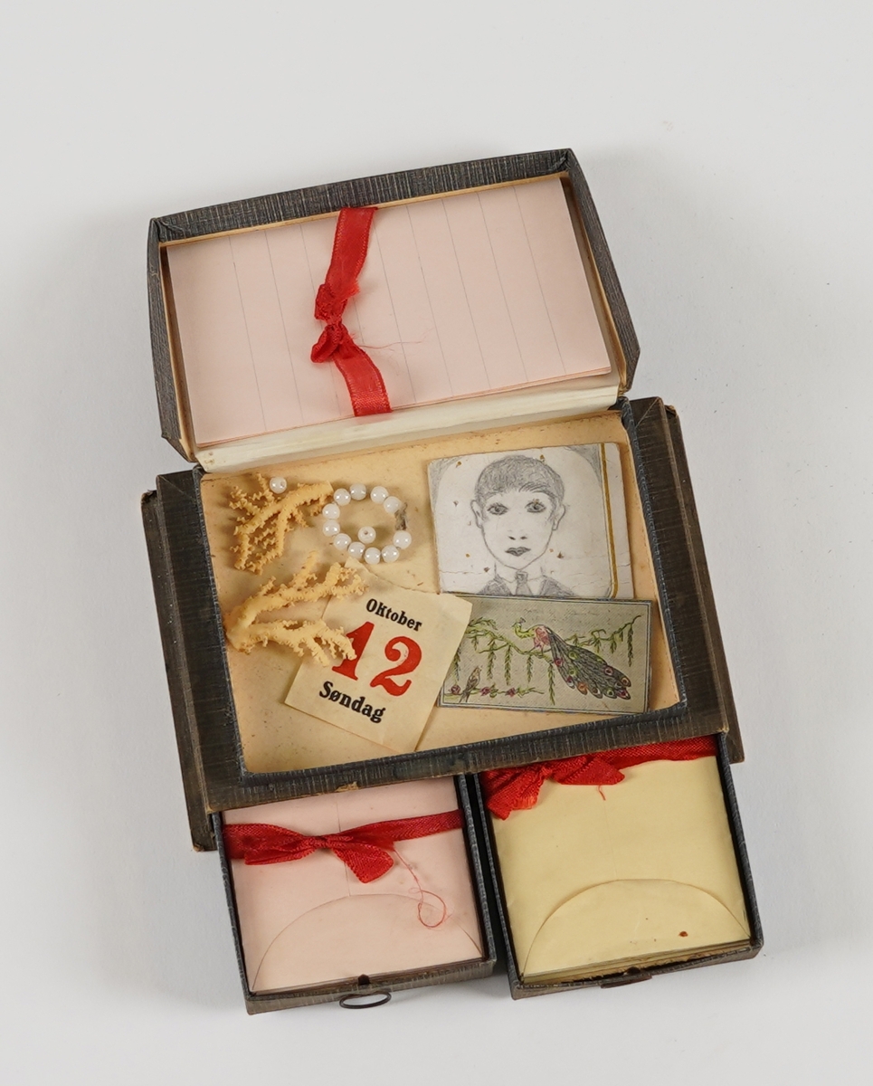 Ei rektangulær øskje med tre rom; to skuffer nedst og ei flat øskje på toppen. I skuffene er det små papirkonvoluttar med raudt band rundt, og i toppen er det små, linjerte ark. I øskja på toppen er det òg diverse små "skattar"; eit teikna portrett, eit utklypt bilete av ein påfugl, eit lite perlearmband (+to lause perler), nokre små korallbitar og ein kalendarlapp (Oktober 12 Søndag). På loket på toppen er det bilete av tre dokkeliknande figurar, og ein liten hund.