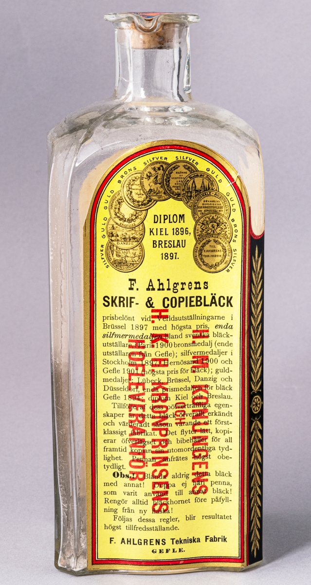 Glasflaska med kork innehållande bläck. Flaskan fyrsidig. Etikett med text "Skrif- & Kopiebläck" på framsidan.
