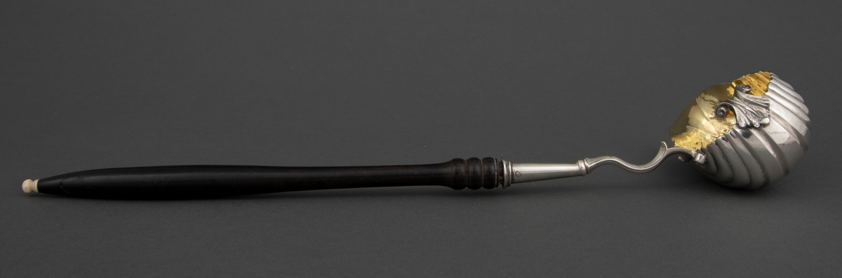 Punsjeøse i sølv med skaft av mørkt løvtre med knapp av ben eller elfenben. Den ovale skålen er innvendig forgylt og har et skjenkenebb på tvers av skaftets retning. Skålen har drevne riller og dekor i lavt relieff.