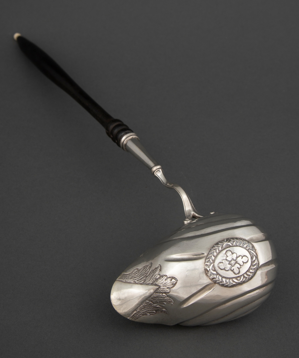 Punsjeøse i sølv med skaft av mørkt løvtre med knapp av ben eller elfenben. Den ovale skålen er innvendig forgylt og har et skjenkenebb på tvers av skaftets retning. Skålen har drevne riller og dekor i lavt relieff.