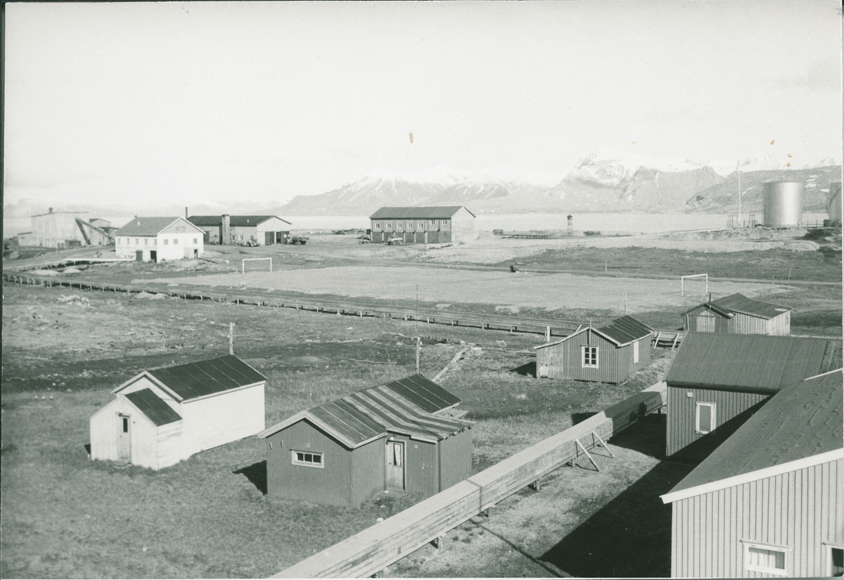 Ny-Ålesund. deler av forsamlingshus, tre familieboliger, fotballplpass, oljetank, flaggstang, garasje, fjøs og mek. verksted. Kongsfjorden og Blomstrand bak.