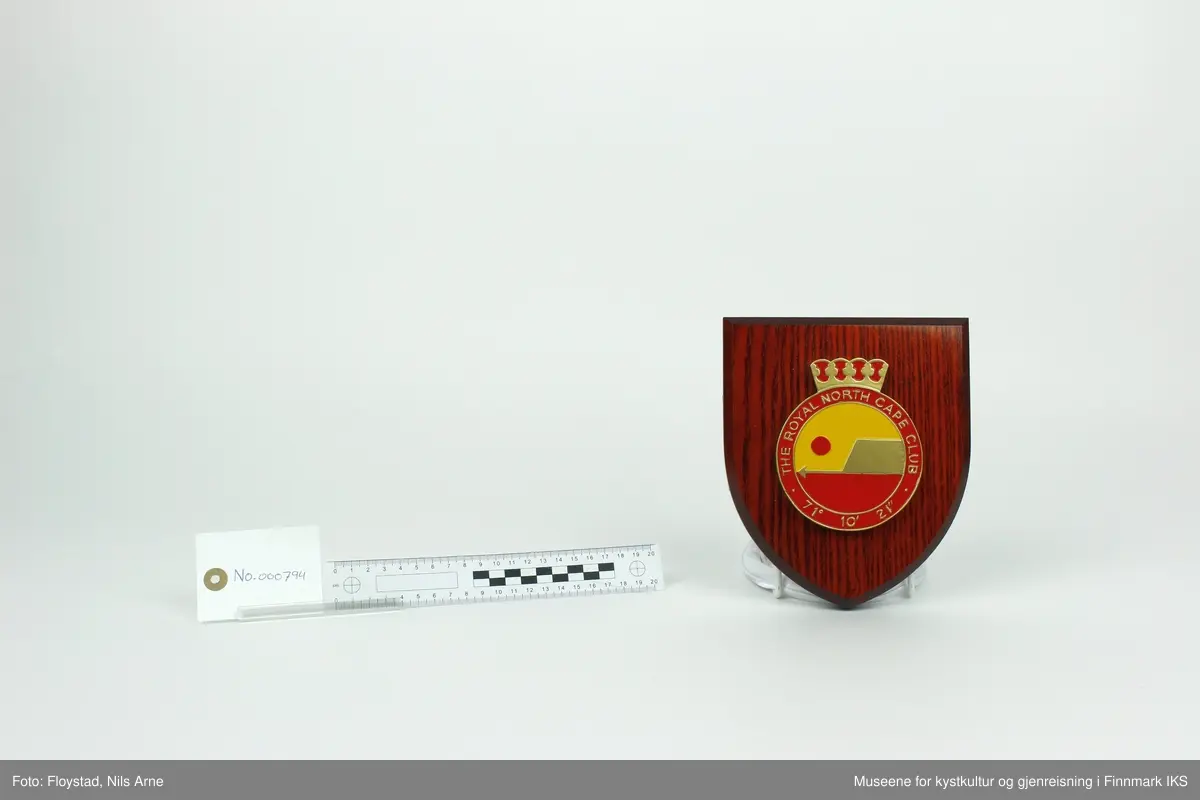 Et våpenskjold dekorert med logoen til The Royal North Cape Club (RNCC). 

Skjoldet har en trekantformet trebase av tresorten ask og er impregnert med mørk beis. Midt på skjoldet er logoen til The Royal North Cape Club limt på. Logoen er også skjært ut av tre og malt i fargene gul, rød og gull. På baksiden er det skjært ut et hull med et feste for oppheng laget av plast. På feste står det "MADE IN GREAT BRITAIN". 

Våpenskjoldet ligger i en blå eske og på lokket og den ene siden er det trykt logoen til produsenten GK Beaulah & Co LTD. På baksiden er det trykt opplysninger om hvilke produkter firmaet lager.