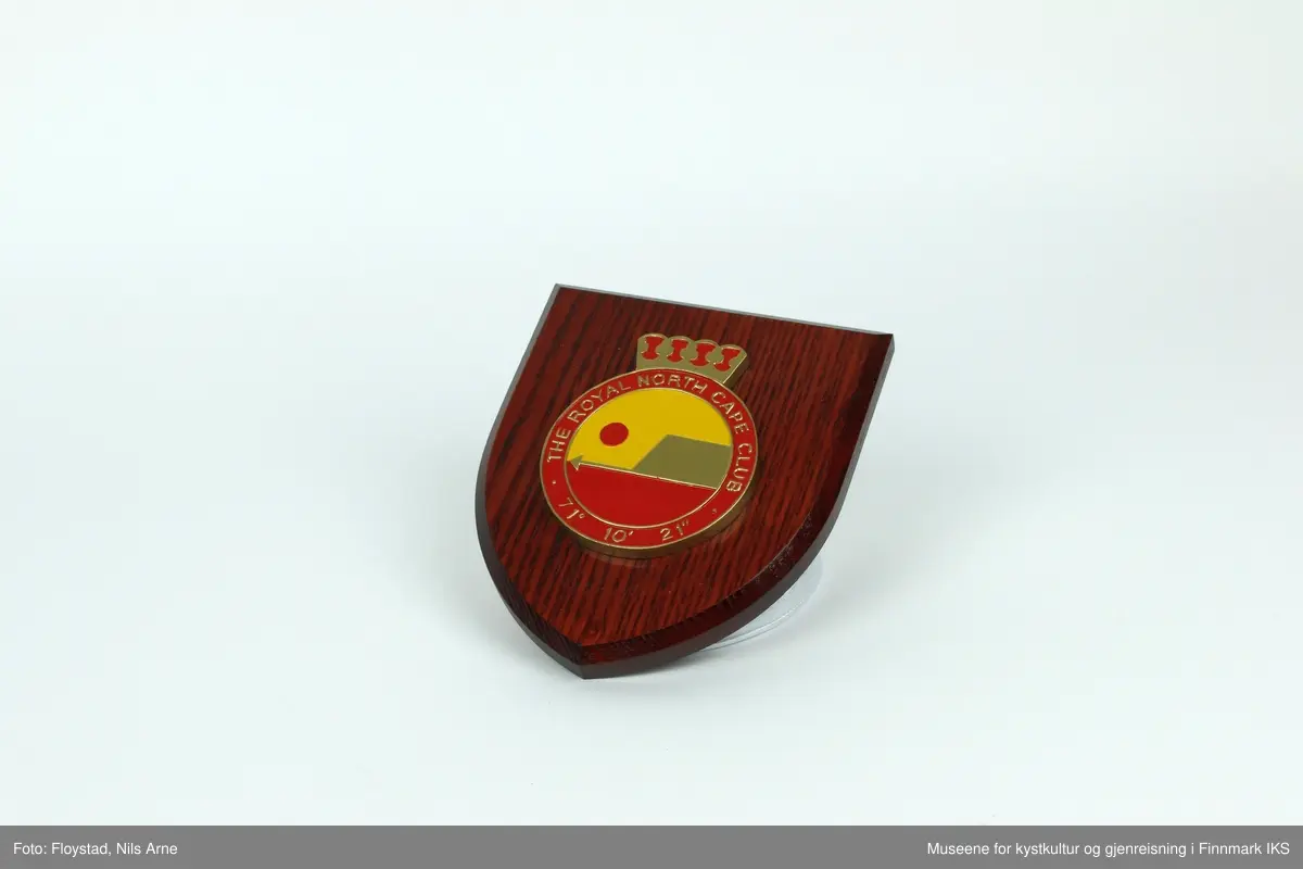Et våpenskjold dekorert med logoen til The Royal North Cape Club (RNCC). 

Skjoldet har en trekantformet trebase av tresorten ask og er impregnert med mørk beis. Midt på skjoldet er logoen til The Royal North Cape Club limt på. Logoen er også skjært ut av tre og malt i fargene gul, rød og gull. På baksiden er det skjært ut et hull med et feste for oppheng laget av plast. På feste står det "MADE IN GREAT BRITAIN". 

Våpenskjoldet ligger i en blå eske og på lokket og den ene siden er det trykt logoen til produsenten GK Beaulah & Co LTD. På baksiden er det trykt opplysninger om hvilke produkter firmaet lager.