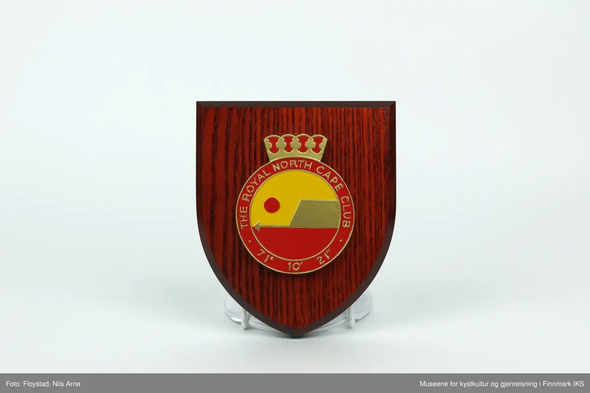 Et våpenskjold dekorert med logoen til The Royal North Cape Club (RNCC). 

Skjoldet har en trekantformet trebase av tresorten ask og er impregnert med mørk beis. Midt på skjoldet er logoen til The Royal North Cape Club limt på. Logoen er også skjært ut av tre og malt i fargene gul, rød og gull. På baksiden er det skjært ut et hull med et feste for oppheng laget av plast. På feste står det "MADE IN GREAT BRITAIN". 

Våpenskjoldet ligger i en blå eske og på lokket og den ene siden er det trykt logoen til produsenten GK Beaulah & Co LTD. På baksiden er det trykt opplysninger om hvilke produkter firmaet lager.