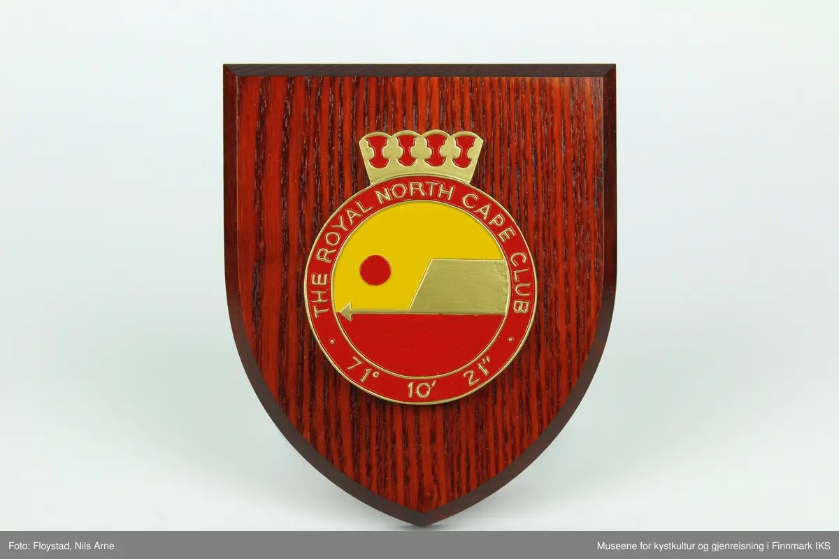 Et våpenskjold dekorert med logoen til The Royal North Cape Club (RNCC). 

Skjoldet har en trekantformet trebase av tresorten ask og er impregnert med mørk beis. Midt på skjoldet er logoen til The Royal North Cape Club limt på. Logoen er også skjært ut av tre og malt i fargene gul, rød og gull. På baksiden er det skjært ut et hull med et feste for oppheng laget av plast. På feste står det "MADE IN GREAT BRITAIN". 

Våpenskjoldet ligger i en blå eske og på lokket og den ene siden er det trykt logoen til produsenten GK Beaulah & Co LTD. På baksiden er det trykt opplysninger om hvilke produkter firmaet lager.