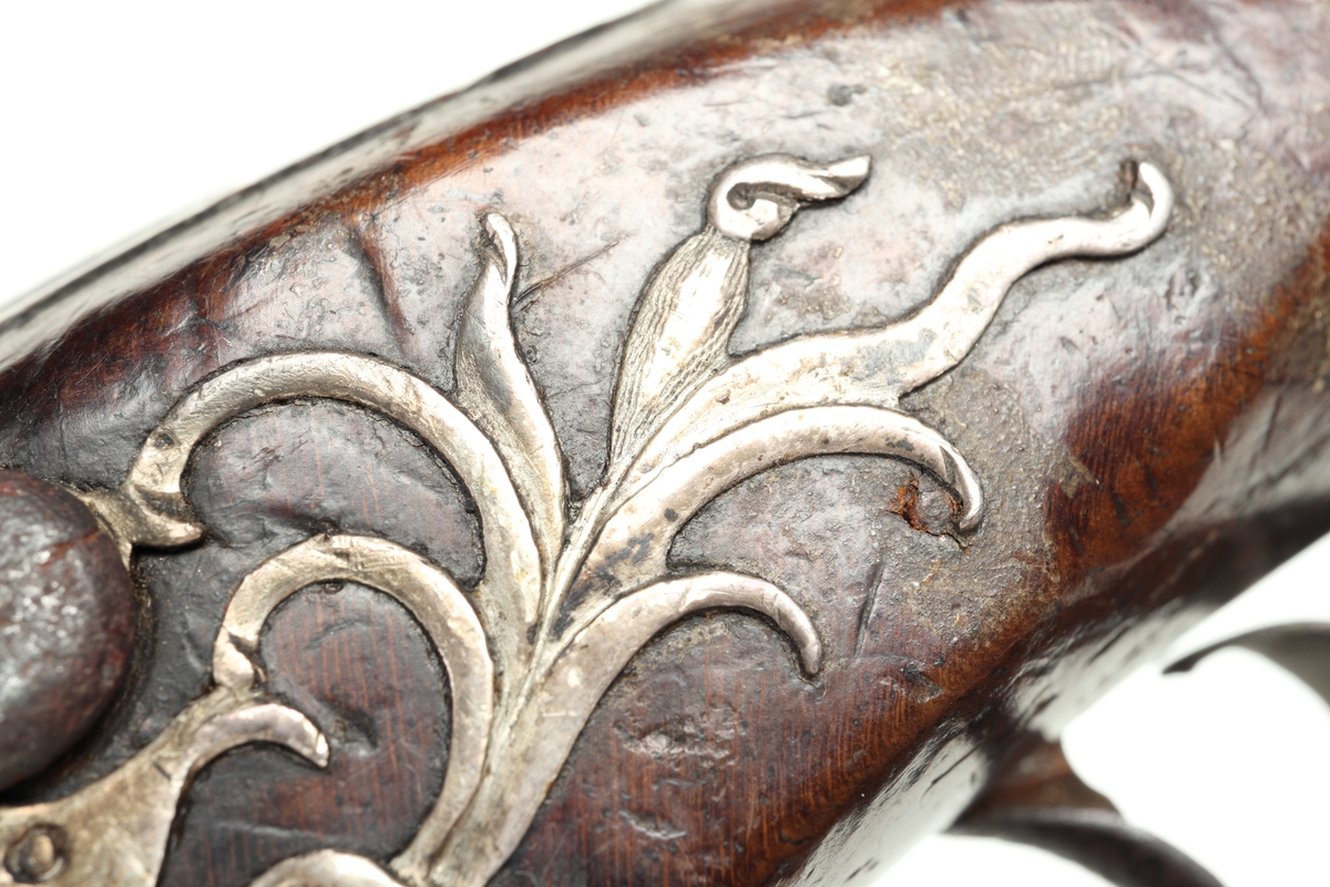 Flintlåspistol med helstock och lås i stål i s.k. Centuckystil. Pipan är gjord i mässing, och kolvkappa, varbygel, sidobleck, tumplåt och rörkor i silver. Pipan är märkt "ST-965", samt två kontrollstämplar från London, och som är åttakantig i formen, men övergår i cylindrisk form ut mot mynningen. Kolvkappan har silverstämplar från Birmingham, varbygeln likaså. Sidoblecket är utformat som blomsterslingor, och kolvkappan som en maskaron. Varbygeln är ornamenterad med en blomma i en oval. Längst fram är den utformad som ett ekollon. Tumplåten är märkt med "EC". Laddstock finns under framstocken.
Inskrivet i huvudkatalogen 1974.