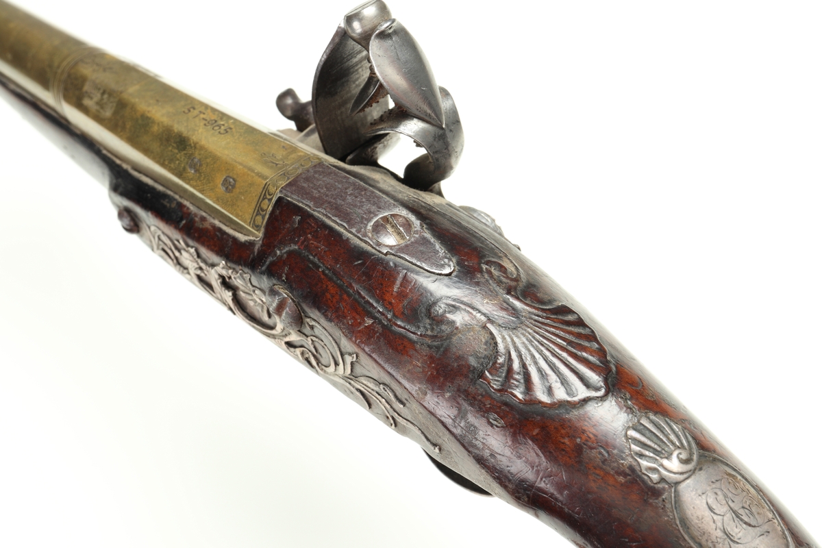 Flintlåspistol med helstock och lås i stål i s.k. Centuckystil. Pipan är gjord i mässing, och kolvkappa, varbygel, sidobleck, tumplåt och rörkor i silver. Pipan är märkt "ST-965", samt två kontrollstämplar från London, och som är åttakantig i formen, men övergår i cylindrisk form ut mot mynningen. Kolvkappan har silverstämplar från Birmingham, varbygeln likaså. Sidoblecket är utformat som blomsterslingor, och kolvkappan som en maskaron. Varbygeln är ornamenterad med en blomma i en oval. Längst fram är den utformad som ett ekollon. Tumplåten är märkt med "EC". Laddstock finns under framstocken.
Inskrivet i huvudkatalogen 1974.