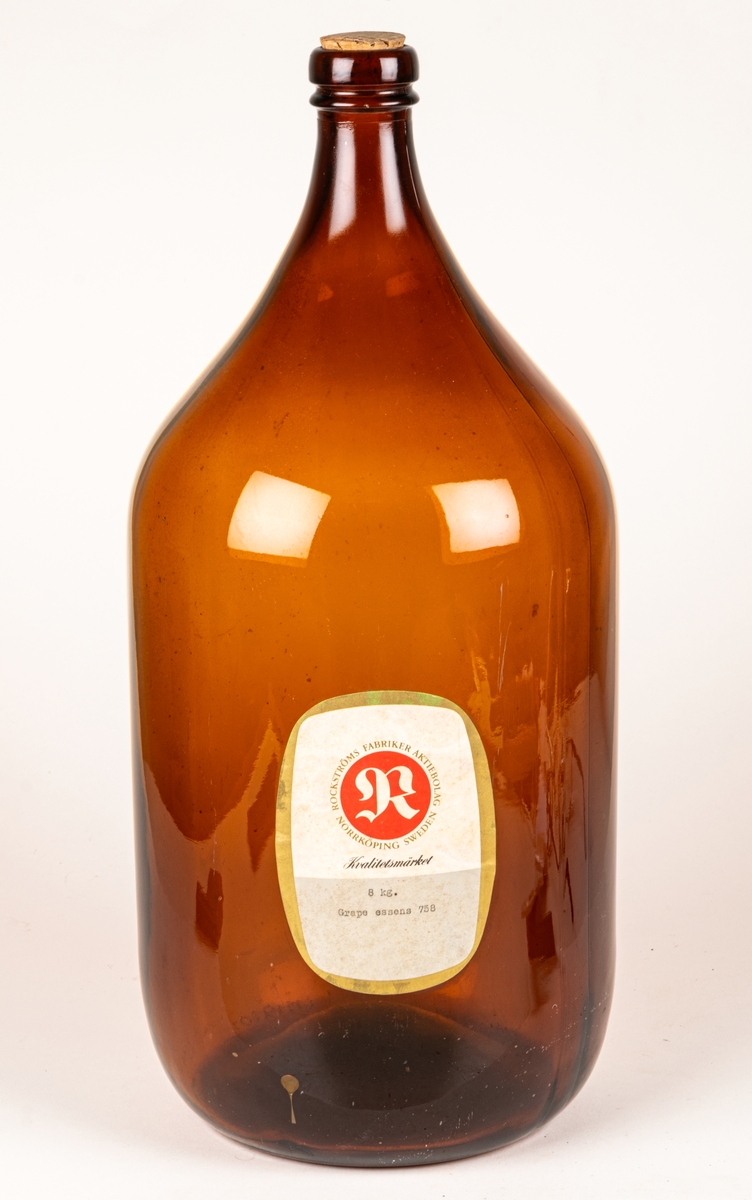 Brun glasflaska. Rektangulär grå och vit etikett med guldkant. Rött sigill med vitt "R", text runt sigillet: "ROCKSTRÖMS FABRIKER AKTIEBOLAG NORRKÖPING SWEDEN" samt "KVALITETSMÄRKET 8 KG GRAPE ESSENS 758."