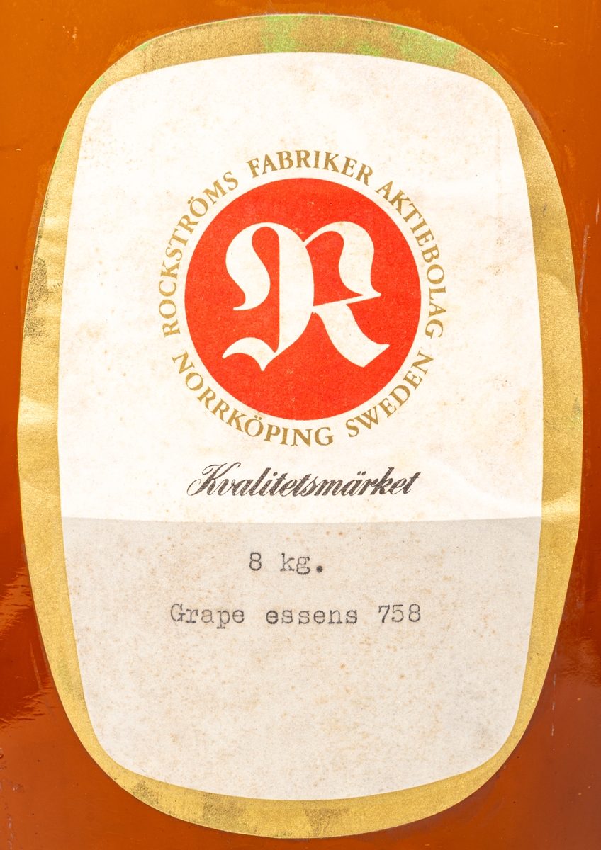 Brun glasflaska. Rektangulär grå och vit etikett med guldkant. Rött sigill med vitt "R", text runt sigillet: "ROCKSTRÖMS FABRIKER AKTIEBOLAG NORRKÖPING SWEDEN" samt "KVALITETSMÄRKET 8 KG GRAPE ESSENS 758."