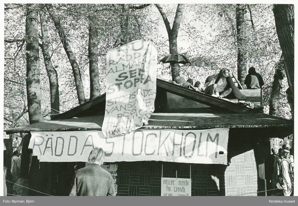 Almarna, almstriden i Kungsträdgården, Stockholm, 12-13 maj 1971, då ett antal miljövänner genom att klättra upp i de hundraåriga almarna lyckades hindra att träden sågades ned för att ge plats till en planerad tunnelbaneuppgång. Banderoller och plakat med text "RÄDDA STOCKHOLM" "NU RÄDDA ALMARNA SEN STOPPA STORSTADSVANSINNET I REGI ...".