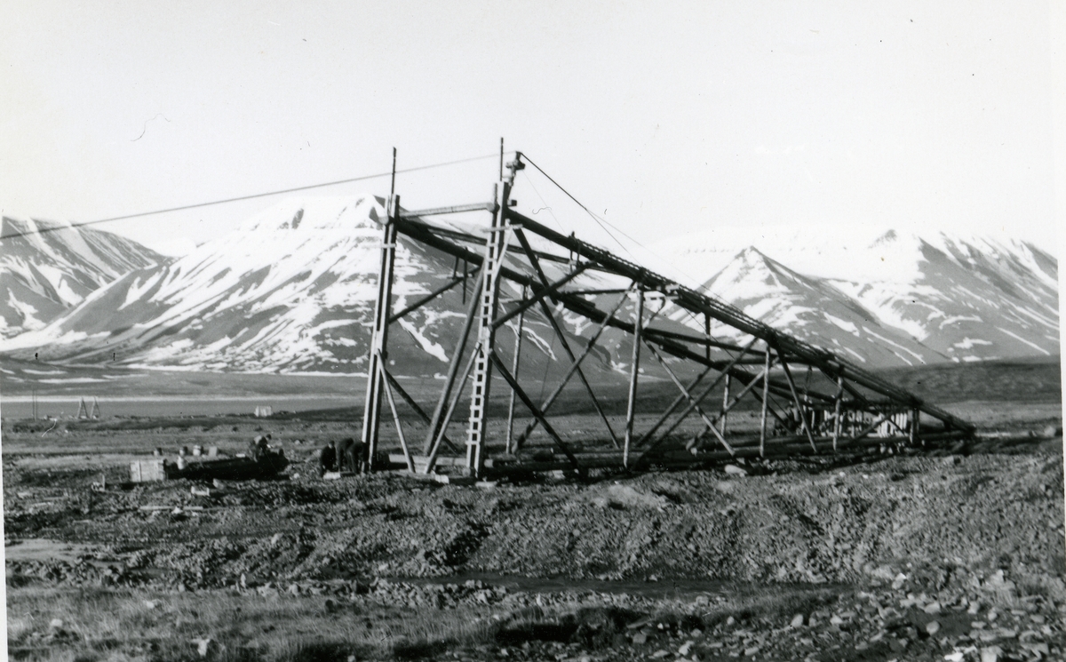 Reising av taubanebukk i Longyeardalen.Taubanetrasèen til gruve 5 bygges sommeren 1959. Dette var den høyeste bukken på banen til Gruve 5. Den ble reist ved hjelp avto bulldosere med wire og spill. En reiste og en holdt igjen. Så ble bukken senket ned ved hjelp av jekker. Bulldoserkjørerne var Tor Kristoffersen og Kåre G. Pedersen. Samme motiv som SVF 13083, tatt av Erling Nødtvedt. Kan være en kopi. 2 eks.