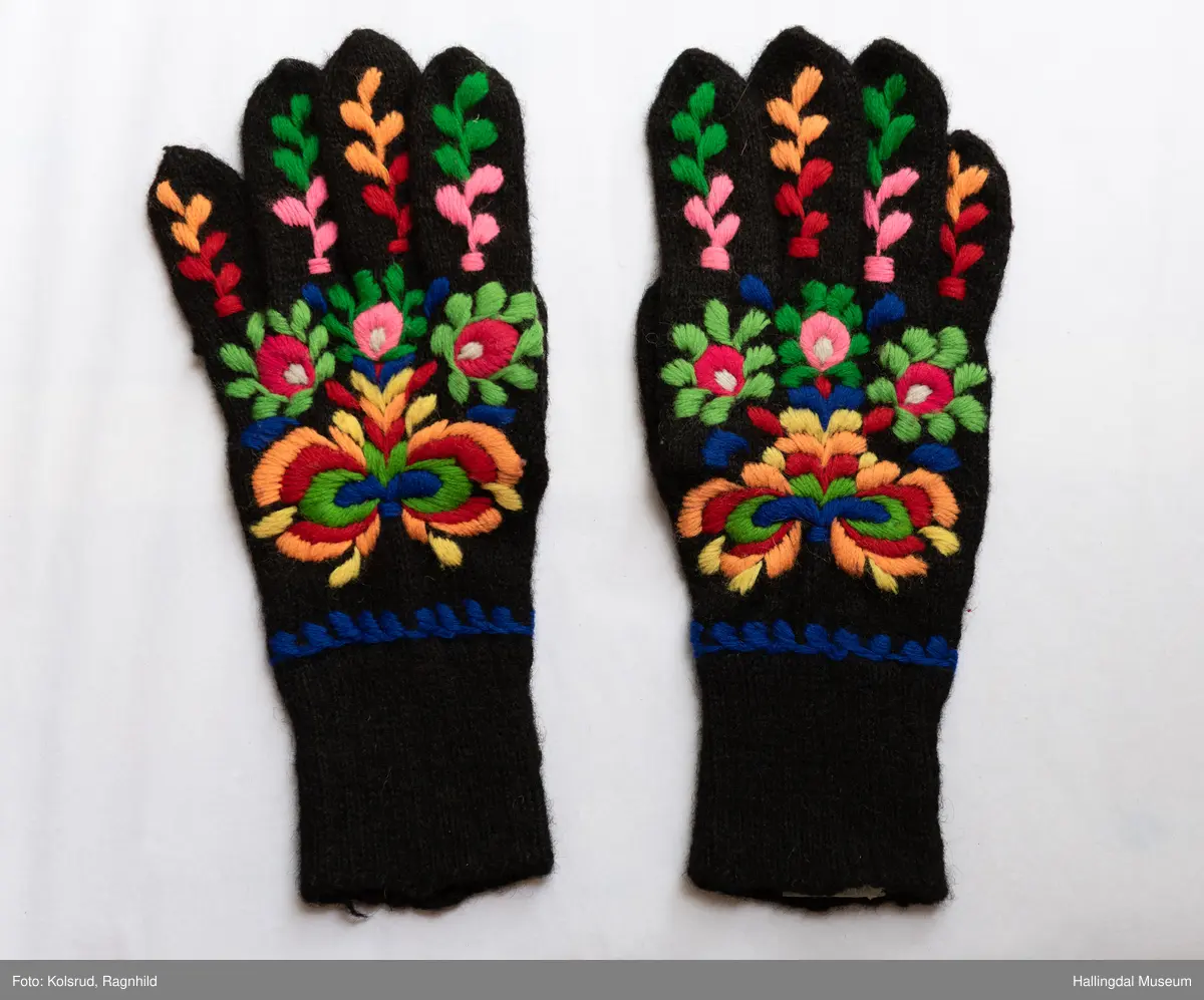 Heimestrikka i svart ull og rikt brodert med plattsaumbroderi på heile oversida. Blå bord rundt handleddet. Nyansar i raudt, grønt og gult, samt kvitt og blått. Vrangborden er 8 cm lang, og strikka 2 rett og 1 vrang. 