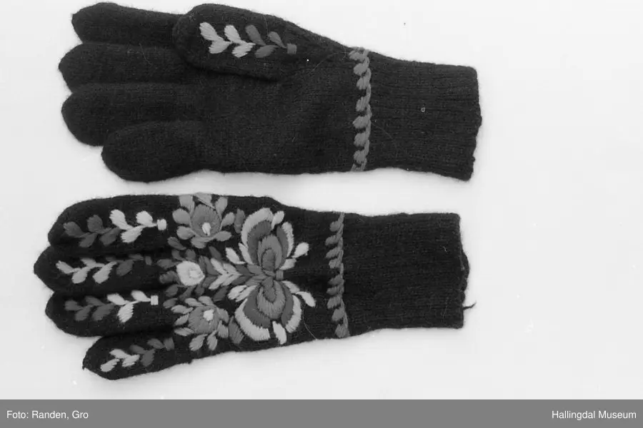 Heimestrikka i svart ull og rikt brodert med plattsaumbroderi på heile oversida. Blå bord rundt handleddet. Nyansar i raudt, grønt og gult, samt kvitt og blått. Vrangborden er 8 cm lang, og strikka 2 rett og 1 vrang. 