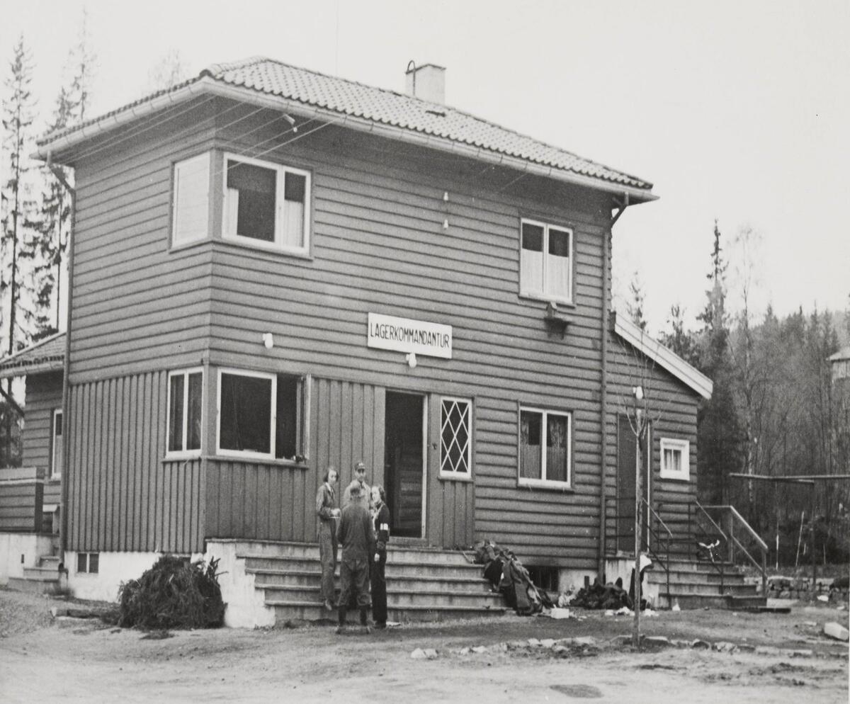 Fotografi av Kommandantur-bygningen utenfor. På fremsiden av bygget er det et skilt hvor det står: Lagerkommandantur. Fire mennesker står foran bygget.