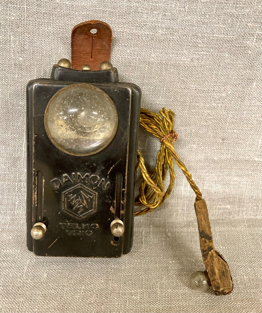 Lommelykt med ekstra uttak, med en ledning som gir strøm til en liten lyspære montert på et trehåndtak. Festereim i lær med knappehull.