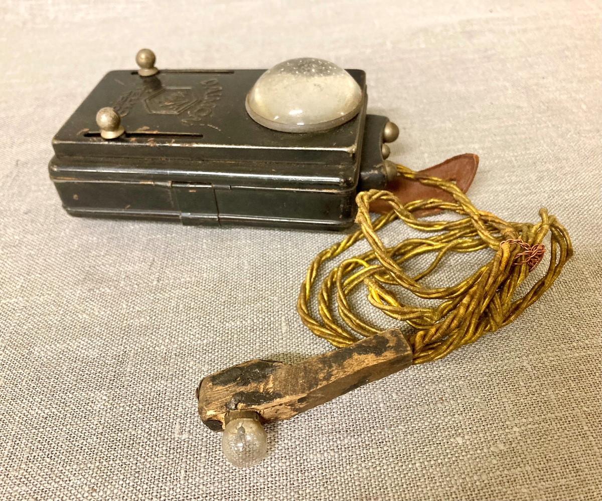 Lommelykt med ekstra uttak, med en ledning som gir strøm til en liten lyspære montert på et trehåndtak. Festereim i lær med knappehull.