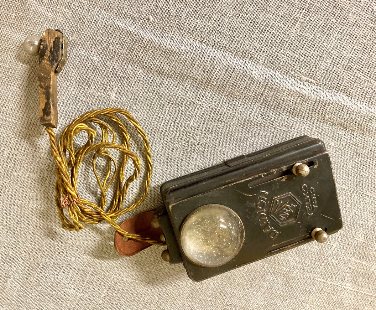 Lommelykt med ekstra uttak, med en ledning som gir strøm til en liten lyspære montert på et trehåndtak. Festereim i lær med knappehull.