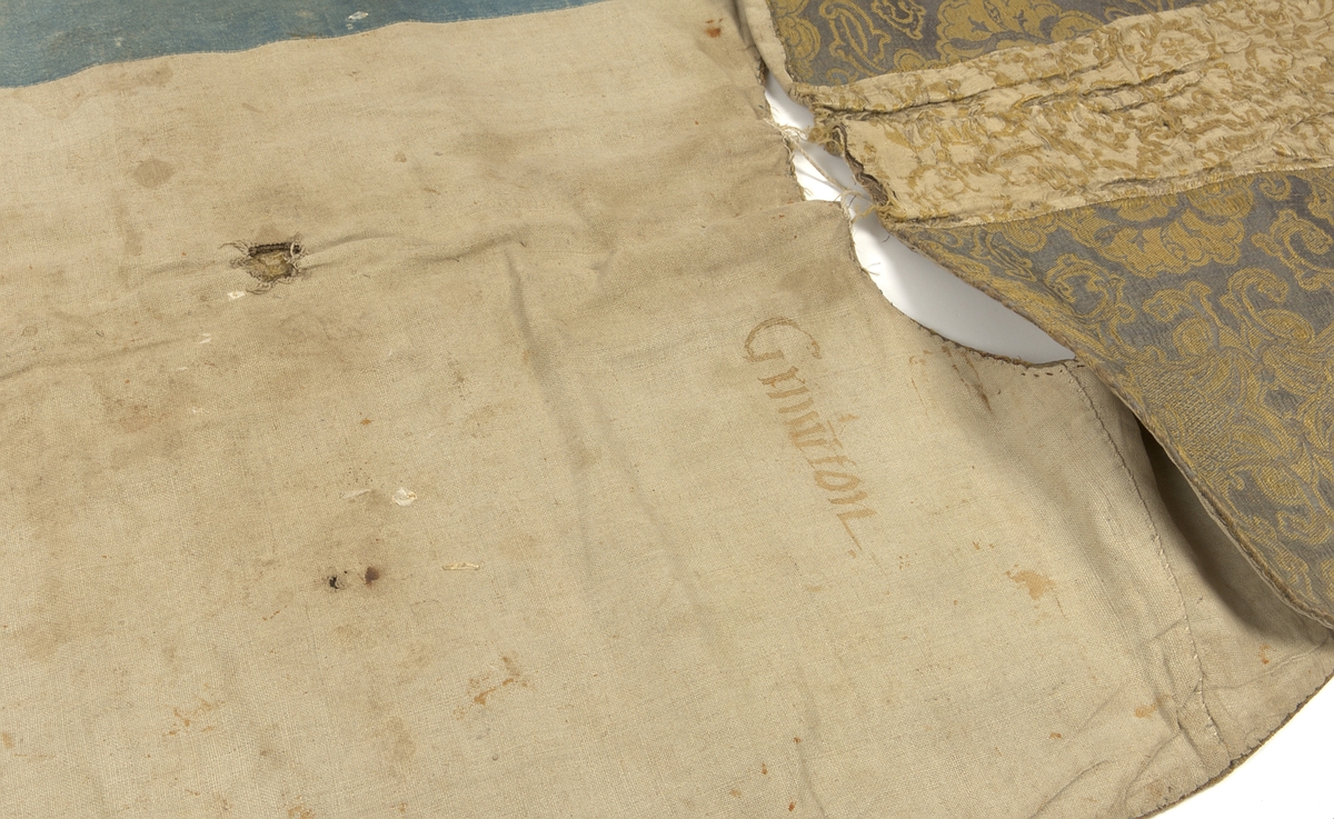 Mässhake. Sidendamast, varp violett med gult samt silver i inslag. Korset i vitt och gult med silverinslag.Beskrivning av tygerna: Sidostycken: Renässans-barockstil. Två varpar: gul, gles varp, röd tät varp 4 skafts balavia. 3 inslagssystem: grovt lininslag, gult silke, metall två trådar tillsammans. Röda varp flotteringar bildar figurernas konturer. 5 skafts satin?Korsfält: Gult och vitt silke, metalltråd, lampasbindning, botten tuskaft, mönster 3 skafts kypert? Vävt varannat gult och metall, varannat vit silke.Foder av olika linne i olika färger. /Lotti 1986