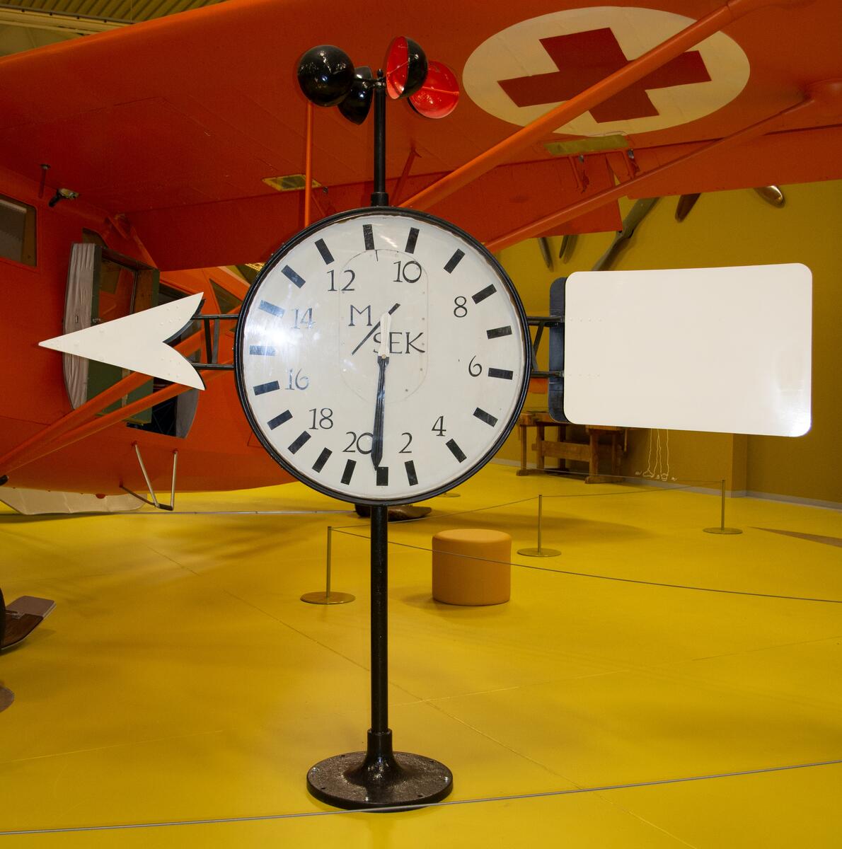 Vindmätare, tillverkad i svart metall med vitmålad vindpekare. Vit urtavala som visar vindhastigheten i meter per sekund. Skålkorsanemometer i svart metall, orangeröda kåpor invändigt