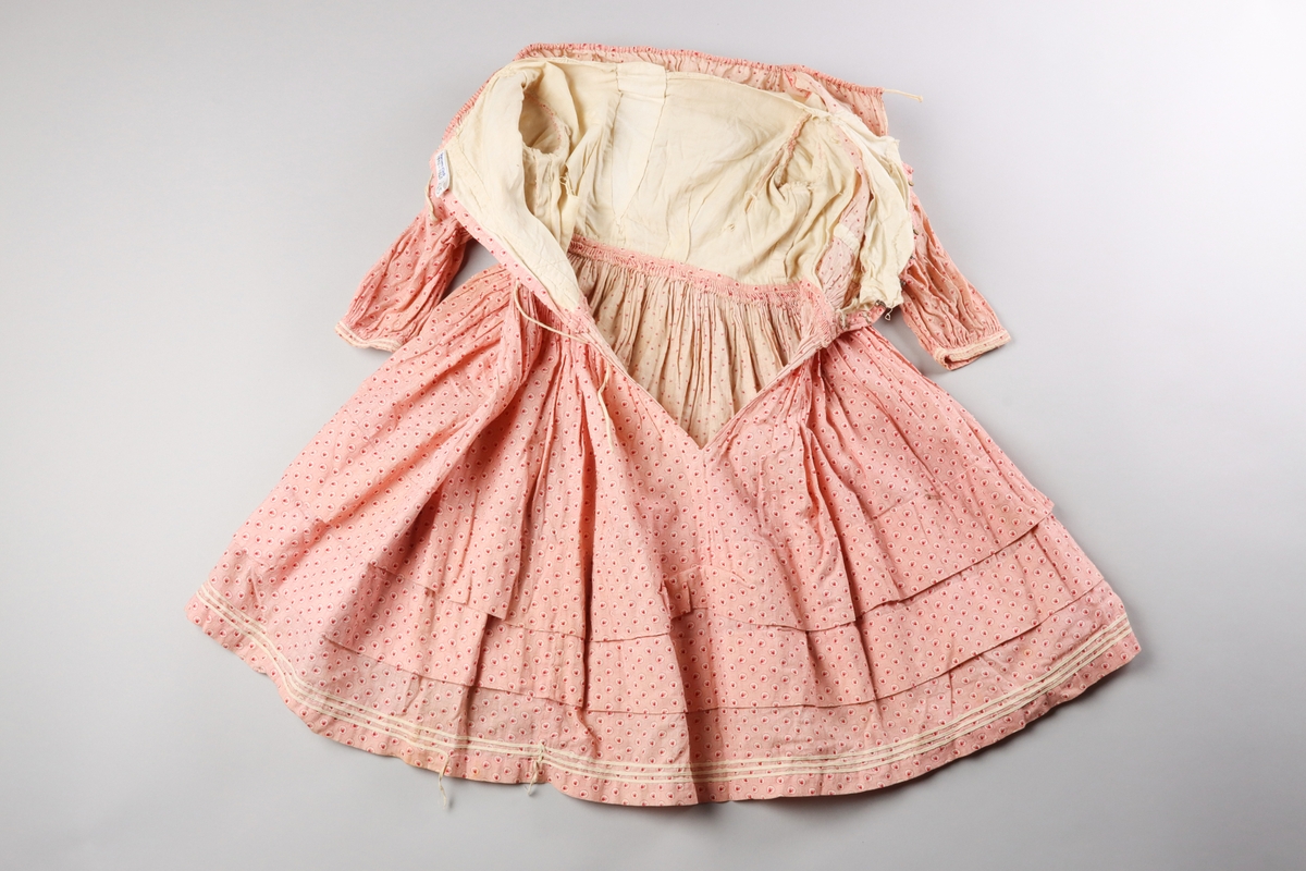 Barnklänning av tät och tunn bomullsväv med tryckt mönster i rosa, rött och vitt. Mönstret föreställer ett litet löv i en cirkel, vilken upprepas som täta prickar mot en rosa botten.

Halsringningen är vid och har en dragsko för reglering. Klänningens liv är rynkat i partier mot midjan. På insidan är ett band påsytt ca 4 cm upp från midjan för att samla ihop rynkorna både på fram och bakstycken. Klänningens liv är fodrat med oblekt bomullsväv som har en isättning i form av en kil mitt fram, troligen för att öka storleken. Klänningens livfoder stängs med hyskor och hakar i mässingstråd och järntråd i en knäppning mitt bak. Knäppningen döljs av de samlade vecken på livets baksida. Veckens band och halsringningens band knyts bak.

Ärmarna är rynkade i en puff närmast axeln. Den del av ärmen har ett orynkat slätt foder av oblekt bomullstyg. Puffärmen har en egen ärmlinning och den längre ärmen med mindre omfång är fäst innuti den. Den längre ärmen är inte fodrad. Det verkar som om den delen av ärmen kan sprättas av för att ändra klänningen till kortärmad. Den nedre ärmen är samlad mot en ärmlinning dekorerad med två parallella vita flätade bomullsband.

Klänningens kjol är veckad med parallella rynkor mot midjan och varje rynka är fäst mot livet med ett stygn. Kjolen öppnas med ett sprund mitt bak. Kjolen har två horisontella veck. Det övre är 6,5 cm djupt och det undre 5,6 cm djupt. Vecken ger kjolens vidd viss stadga, men kan också ha använts för att reglera längden. Kjolens nedre kant är uppvikt och fållad med fållstygn. Längs nederkanten löper tre parallella vita flätade bomulsband.

Hela plagget är handsytt med exempel på flera olika typer av stygn och sömnadstekniker. Fodret i livet har flera revor vilket möjliggör studier av insidan.