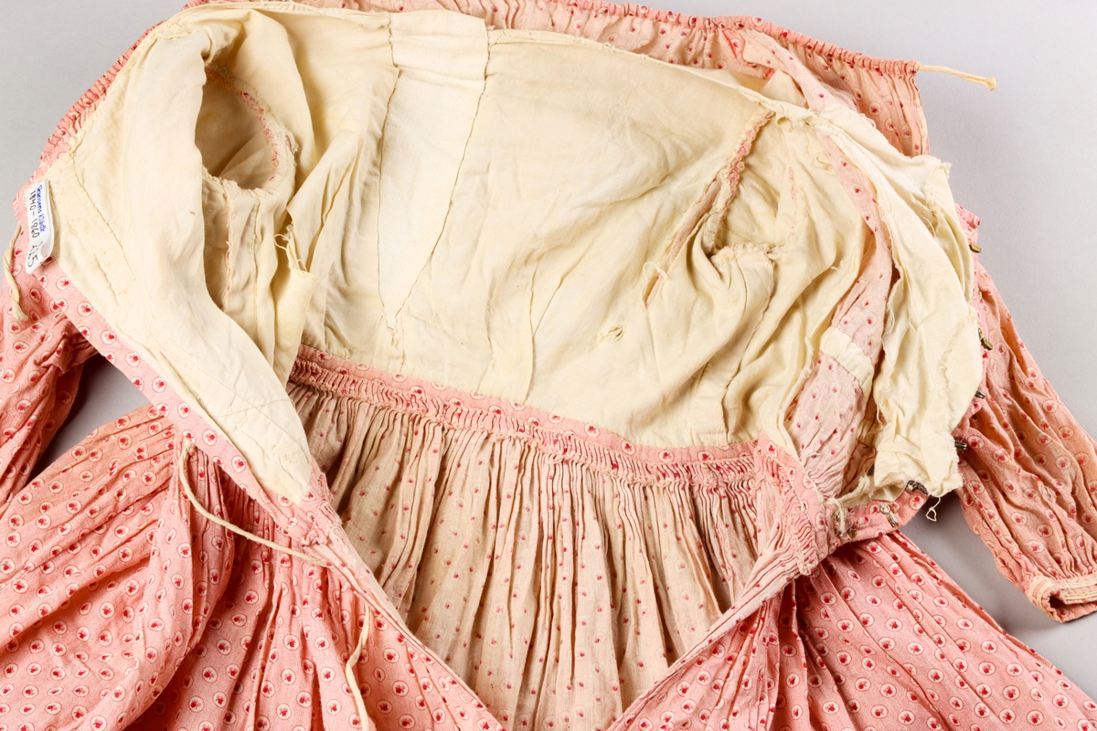 Barnklänning av tät och tunn bomullsväv med tryckt mönster i rosa, rött och vitt. Mönstret föreställer ett litet löv i en cirkel, vilken upprepas som täta prickar mot en rosa botten.

Halsringningen är vid och har en dragsko för reglering. Klänningens liv är rynkat i partier mot midjan. På insidan är ett band påsytt ca 4 cm upp från midjan för att samla ihop rynkorna både på fram och bakstycken. Klänningens liv är fodrat med oblekt bomullsväv som har en isättning i form av en kil mitt fram, troligen för att öka storleken. Klänningens livfoder stängs med hyskor och hakar i mässingstråd och järntråd i en knäppning mitt bak. Knäppningen döljs av de samlade vecken på livets baksida. Veckens band och halsringningens band knyts bak.

Ärmarna är rynkade i en puff närmast axeln. Den del av ärmen har ett orynkat slätt foder av oblekt bomullstyg. Puffärmen har en egen ärmlinning och den längre ärmen med mindre omfång är fäst innuti den. Den längre ärmen är inte fodrad. Det verkar som om den delen av ärmen kan sprättas av för att ändra klänningen till kortärmad. Den nedre ärmen är samlad mot en ärmlinning dekorerad med två parallella vita flätade bomullsband.

Klänningens kjol är veckad med parallella rynkor mot midjan och varje rynka är fäst mot livet med ett stygn. Kjolen öppnas med ett sprund mitt bak. Kjolen har två horisontella veck. Det övre är 6,5 cm djupt och det undre 5,6 cm djupt. Vecken ger kjolens vidd viss stadga, men kan också ha använts för att reglera längden. Kjolens nedre kant är uppvikt och fållad med fållstygn. Längs nederkanten löper tre parallella vita flätade bomulsband.

Hela plagget är handsytt med exempel på flera olika typer av stygn och sömnadstekniker. Fodret i livet har flera revor vilket möjliggör studier av insidan.