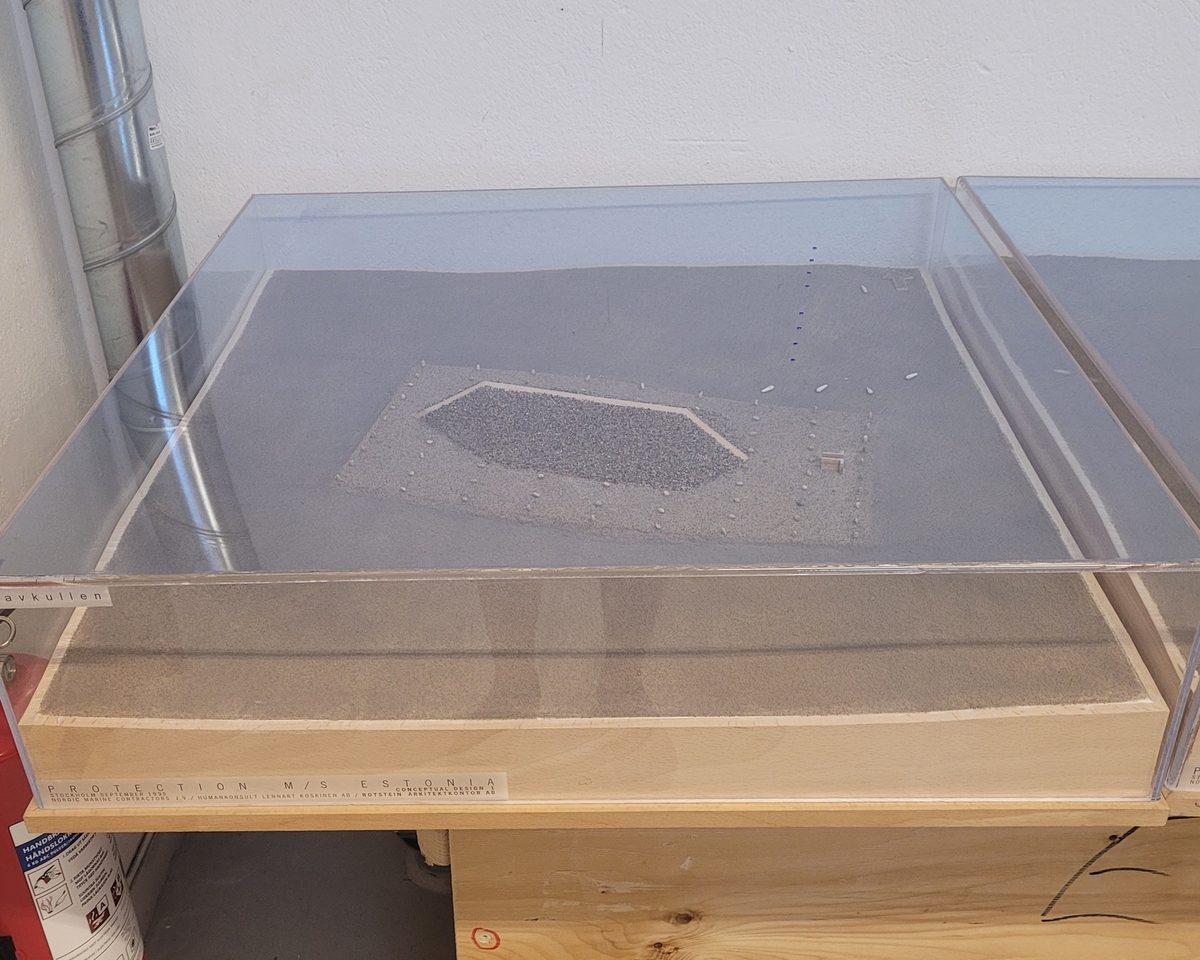 "Gravkullen". Modell till alternativ för den planerade övertäckningen av vraket M/S Estonia. Modellen är utförd i september 1995. Modellen är placerad på träplatta med plexiglashuv. Okänd skala.
8 hål, grupperade i rad 4+4, i plexihuvens tak illustrerar hur bojar var tänkta att märka ut platsen. Modellens "bojar" har dock åkt igenom hålen och ligger på botten.