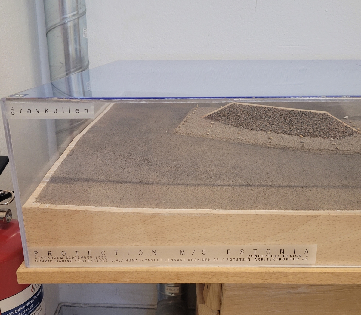 "Gravkullen". Modell till alternativ för den planerade övertäckningen av vraket M/S Estonia. Modellen är utförd i september 1995. Modellen är placerad på träplatta med plexiglashuv. Okänd skala.
8 hål, grupperade i rad 4+4, i plexihuvens tak illustrerar hur bojar var tänkta att märka ut platsen. Modellens "bojar" har dock åkt igenom hålen och ligger på botten.