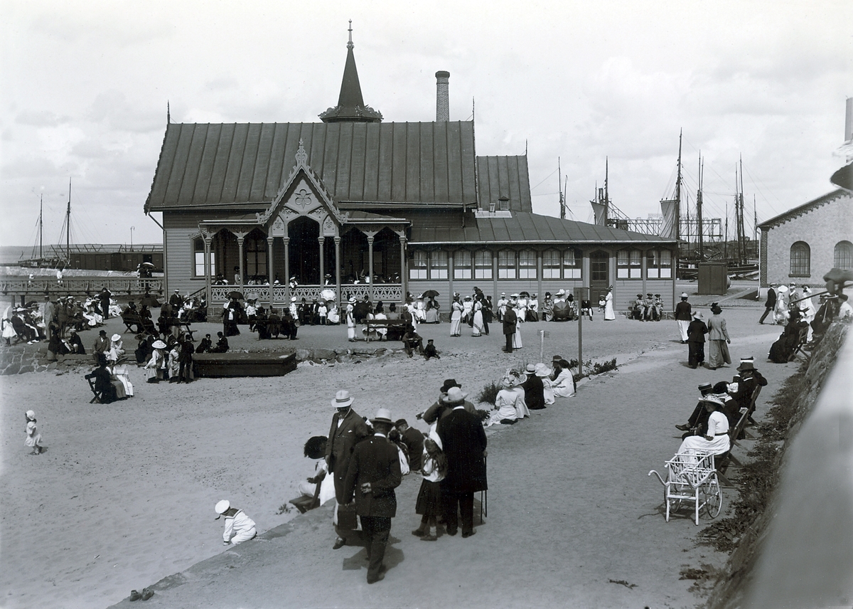 Ur byggmästare Johannes Nilssons fotoalbum från 1914. Avkoppling och umgänge i miljön vid varmbadhuset i Varberg, uppfört 1866. Tre soldater i trekantiga hattar sitter på en bänk till höger vid badhuset. Många damer har solparasoller. I bakgrunden ses master från hamnen och till höger tullhuset, strandpromenaden och uppfarten till fästningen.