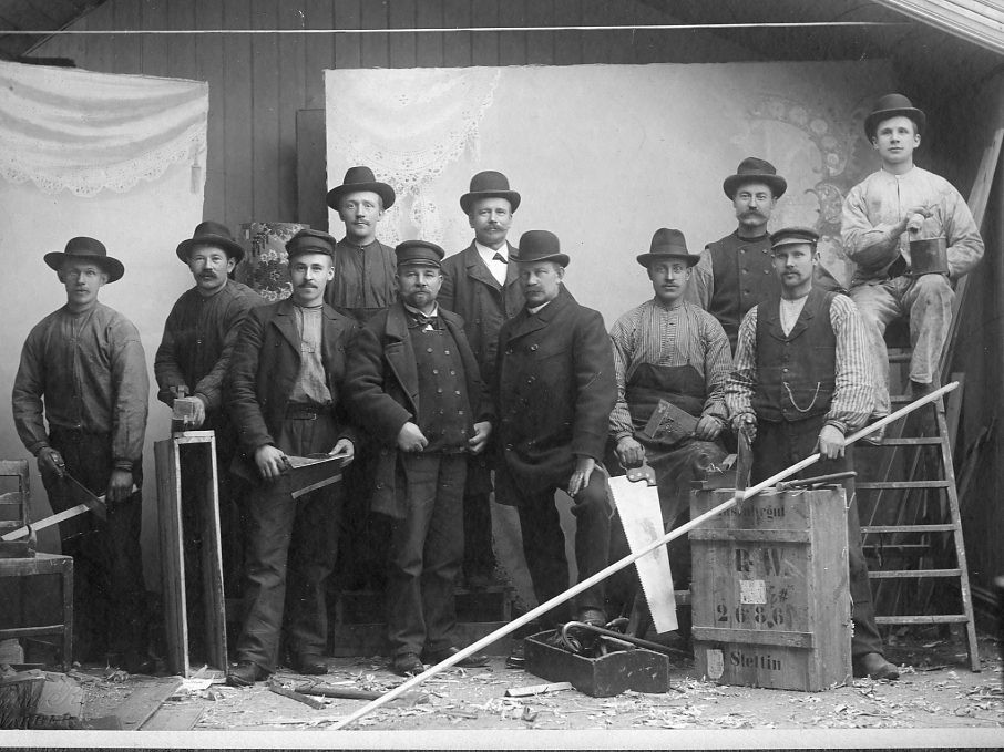 Ur Familjen Nilssons fotosamling. Gruppbild av snickare och målare, anställda hos byggmästare Johannes Nilsson som själv syns iklädd kubb som tredje person i nedre raden från höger.