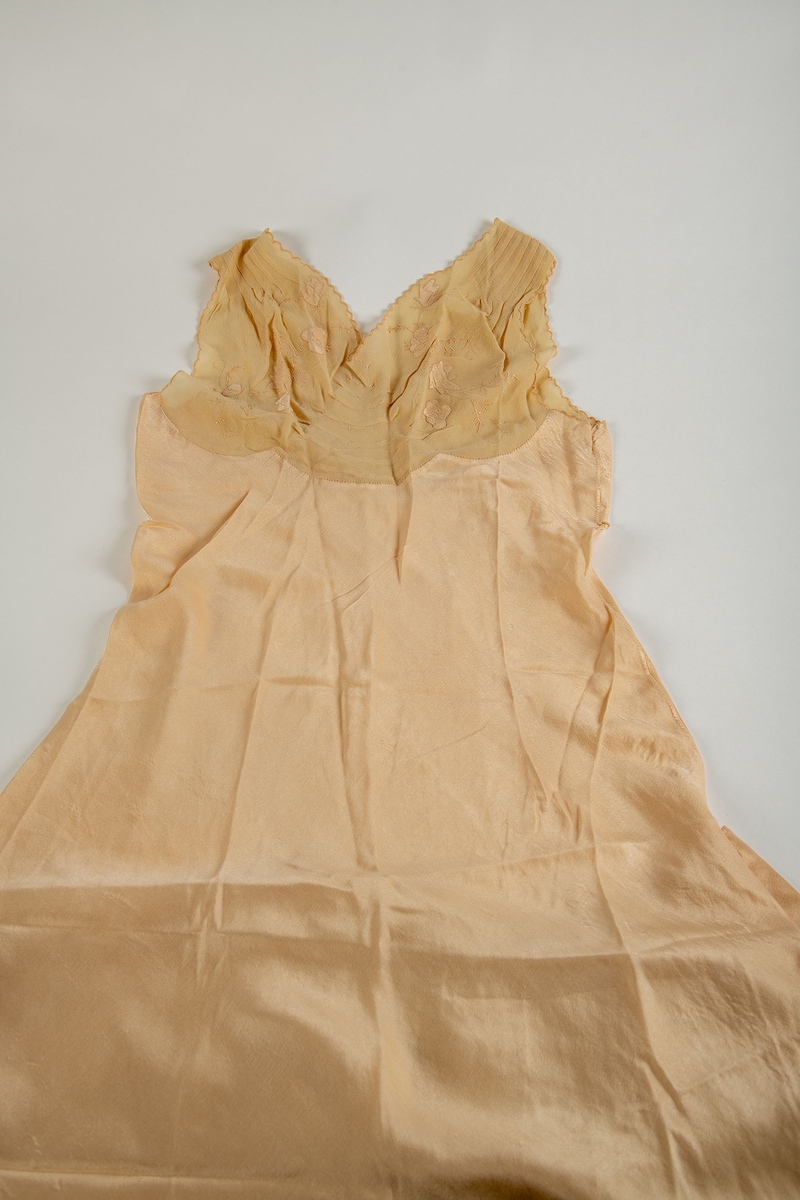 Nattkjole fra Paris, 1930-årene. Silkekjole med lys rosa-beige crépe-de-chine bærestykker.  Tunget bærestykke som i ryggen går over i hverandre i en spiss. 
Foran: legg fra skulderen et kort stykke, buede sømmer på midten, brodert med tynne grener i samme farge som stoffet og med applikerte, firbladete blomster. Tungete kantborder.  Utskrådd skjørt med en kile i hver side.