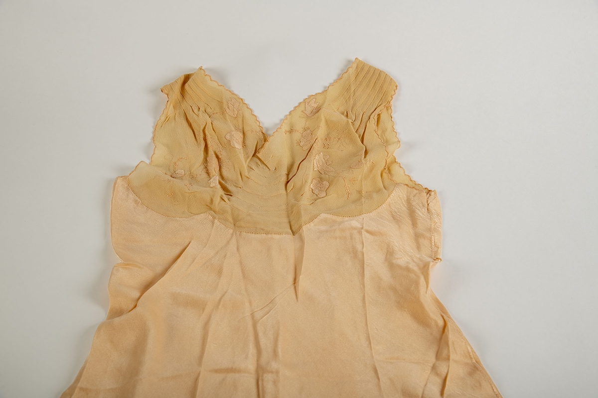 Nattkjole fra Paris, 1930-årene. Silkekjole med lys rosa-beige crépe-de-chine bærestykker.  Tunget bærestykke som i ryggen går over i hverandre i en spiss. 
Foran: legg fra skulderen et kort stykke, buede sømmer på midten, brodert med tynne grener i samme farge som stoffet og med applikerte, firbladete blomster. Tungete kantborder.  Utskrådd skjørt med en kile i hver side.