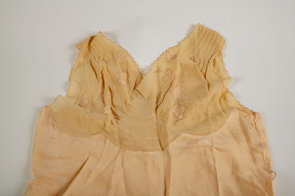 Nattkjole fra Paris, 1930-årene. Silkekjole med lys rosa-beige crépe-de-chine bærestykker.  Tunget bærestykke som i ryggen går over i hverandre i en spiss. 
Foran: legg fra skulderen et kort stykke, buede sømmer på midten, brodert med tynne grener i samme farge som stoffet og med applikerte, firbladete blomster. Tungete kantborder.  Utskrådd skjørt med en kile i hver side.