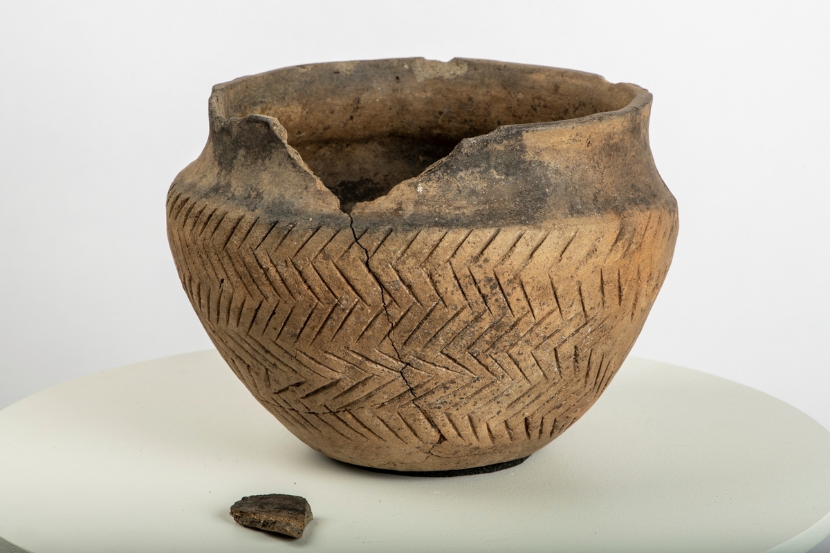 Leirkar av lyst brunlig, temmelig tykt, godt brent gods av form nærmest som Bøe: Keramikk, fig. 11, men med skarpere markert skulder og sterkere skrådd mot bunnen, sml. fig. 10. Randen er rett avskåret, ikke fortykket. Halsen er glatt, sidene er ornert med dypt innrissete streker i fiskebeinsmønster. Orneringen er noe skjødesløst utført, men meget virkningsfull. Noen skår i randen mangler, ellers utmerket bevart.