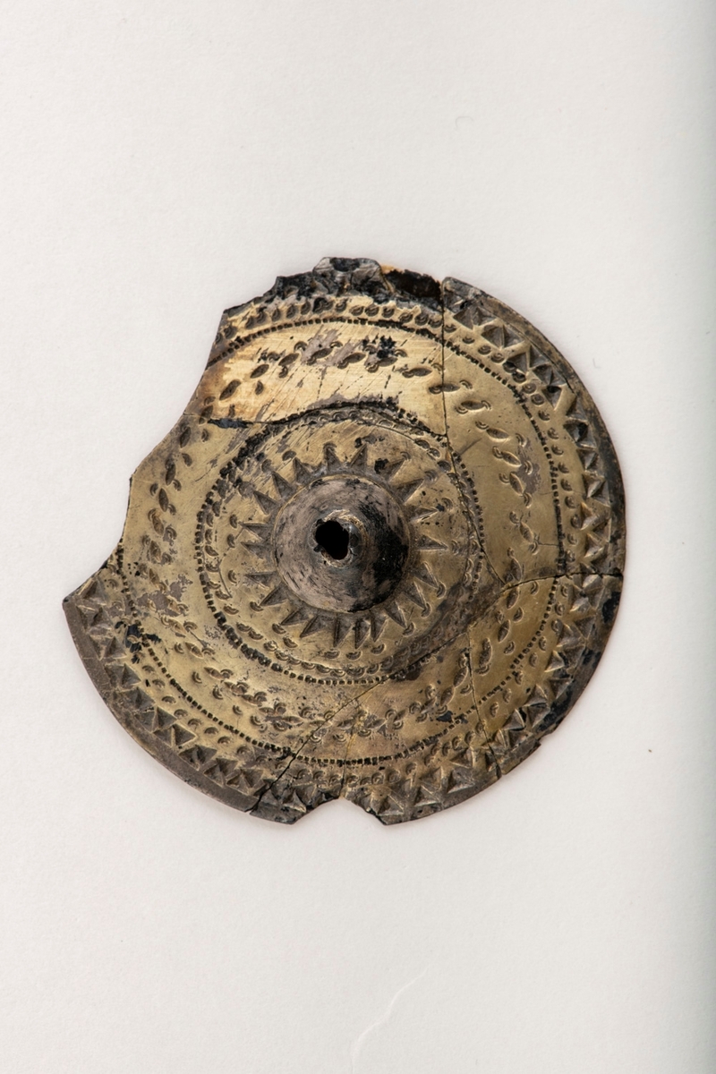 Sirkelrund plate med konisk midtknopp av forgylt sølv av typen [Rygh fig.310]R. 310. Stykket er ornert med konsentriske felter med punsel- og stempelornering, nemlig en dobbelt rad trekanter, så en rekke med små punkter, en tredobbelt rad med noe større punkter og halvmåner på hver side begrenset av en småstiplet linje. Midtstykket er omgitt av en rad sterkt tilspissete trekanter ved hvis toppunkter det finnes små halvmåner. Den ytre begrensning er her en rad små punkter. Stykkets kant er glatt. Foreligger i 5 sammenhørende deler. Noe av kanten mangler. 
