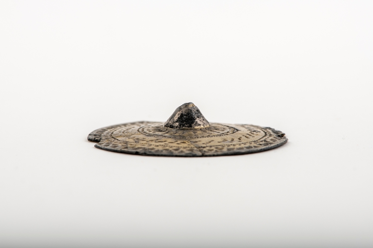 Sirkelrund plate med konisk midtknopp av forgylt sølv av typen [Rygh fig.310]R. 310. Stykket er ornert med konsentriske felter med punsel- og stempelornering, nemlig en dobbelt rad trekanter, så en rekke med små punkter, en tredobbelt rad med noe større punkter og halvmåner på hver side begrenset av en småstiplet linje. Midtstykket er omgitt av en rad sterkt tilspissete trekanter ved hvis toppunkter det finnes små halvmåner. Den ytre begrensning er her en rad små punkter. Stykkets kant er glatt. Foreligger i 5 sammenhørende deler. Noe av kanten mangler. 
