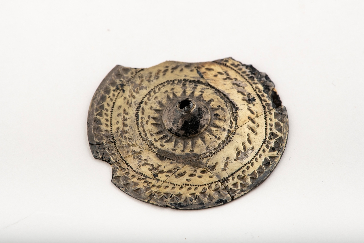 Sirkelrund plate med konisk midtknopp av forgylt sølv av typen [Rygh fig.310]R. 310. Stykket er ornert med konsentriske felter med punsel- og stempelornering, nemlig en dobbelt rad trekanter, så en rekke med små punkter, en tredobbelt rad med noe større punkter og halvmåner på hver side begrenset av en småstiplet linje. Midtstykket er omgitt av en rad sterkt tilspissete trekanter ved hvis toppunkter det finnes små halvmåner. Den ytre begrensning er her en rad små punkter. Stykkets kant er glatt. Foreligger i 5 sammenhørende deler. Noe av kanten mangler. 
