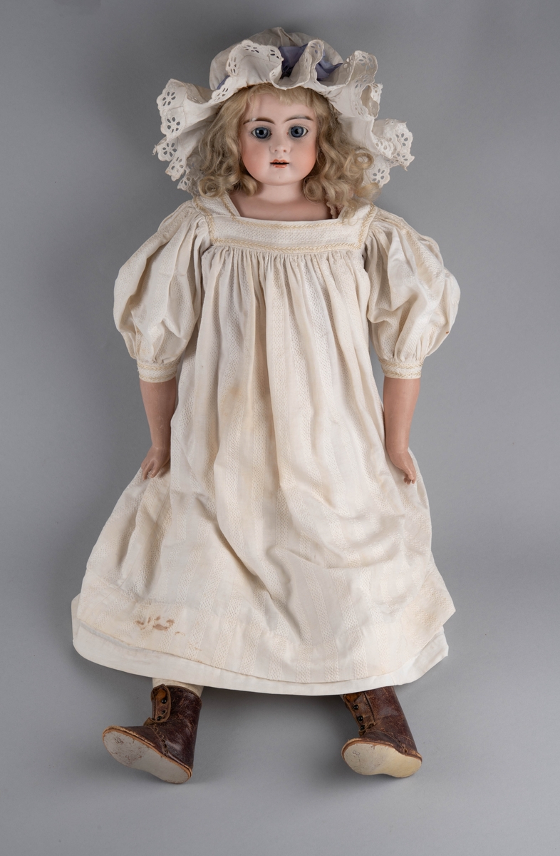 Dukke med porselenshode og underarmer av celluloid. Langt lyst, krøllet hår og blå øyne med bevegelige øyelokk. Stoppet kropp trukket med hanskeskinn, overarmene med tekstil.