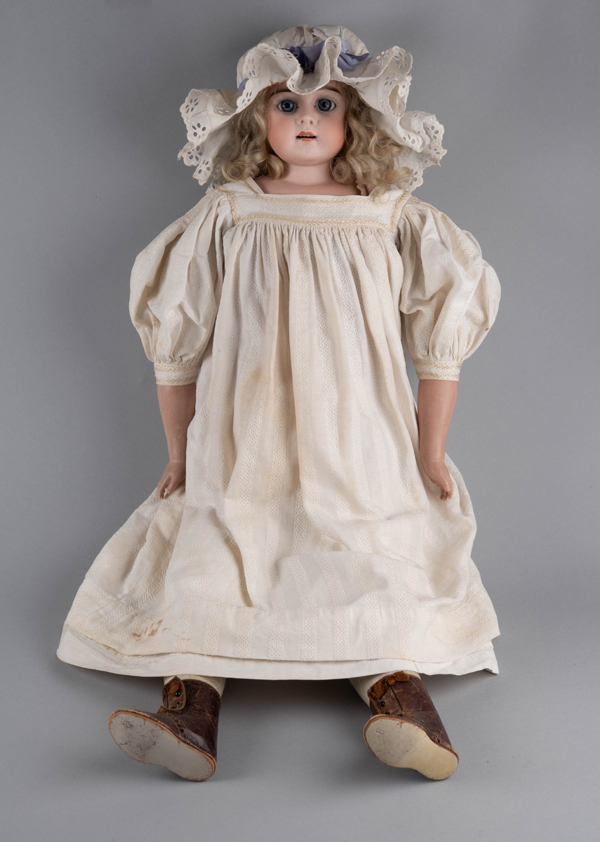 Dukke med porselenshode og underarmer av celluloid. Langt lyst, krøllet hår og blå øyne med bevegelige øyelokk. Stoppet kropp trukket med hanskeskinn, overarmene med tekstil.