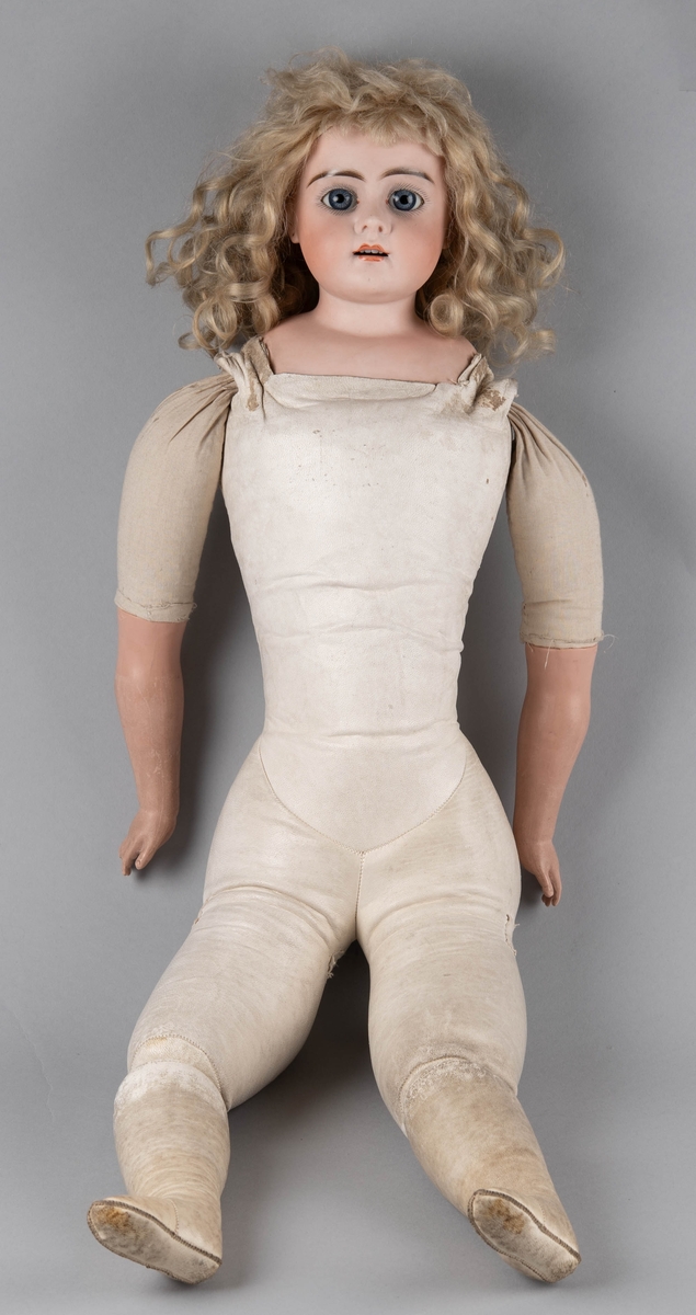 Dukke med porselenshode og underarmer av celluloid. Langt lyst, krøllet hår og blå øyne med bevegelige øyelokk. Stoppet kropp trukket med hanskeskinn, overarmene med tekstil.