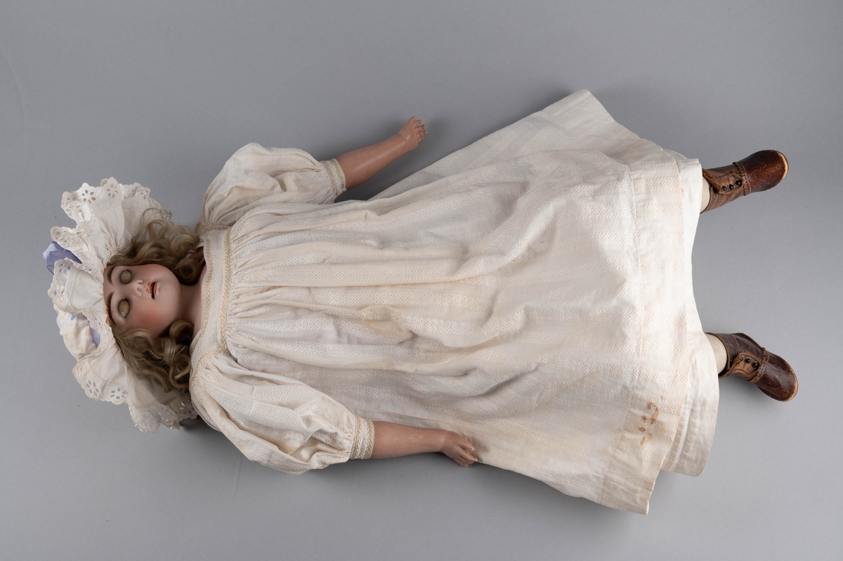 Dukke med porselenshode og underarmer av celluloid. Langt lyst, krøllet hår og blå øyne med bevegelige øyelokk. Stoppet kropp trukket med hanskeskinn, overarmene med tekstil.