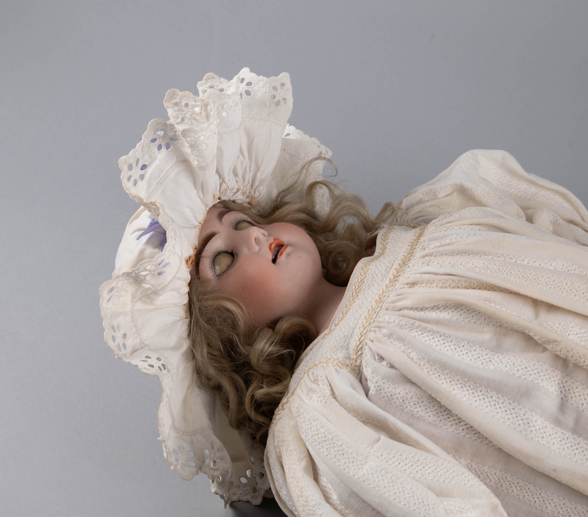 Dukke med porselenshode og underarmer av celluloid. Langt lyst, krøllet hår og blå øyne med bevegelige øyelokk. Stoppet kropp trukket med hanskeskinn, overarmene med tekstil.