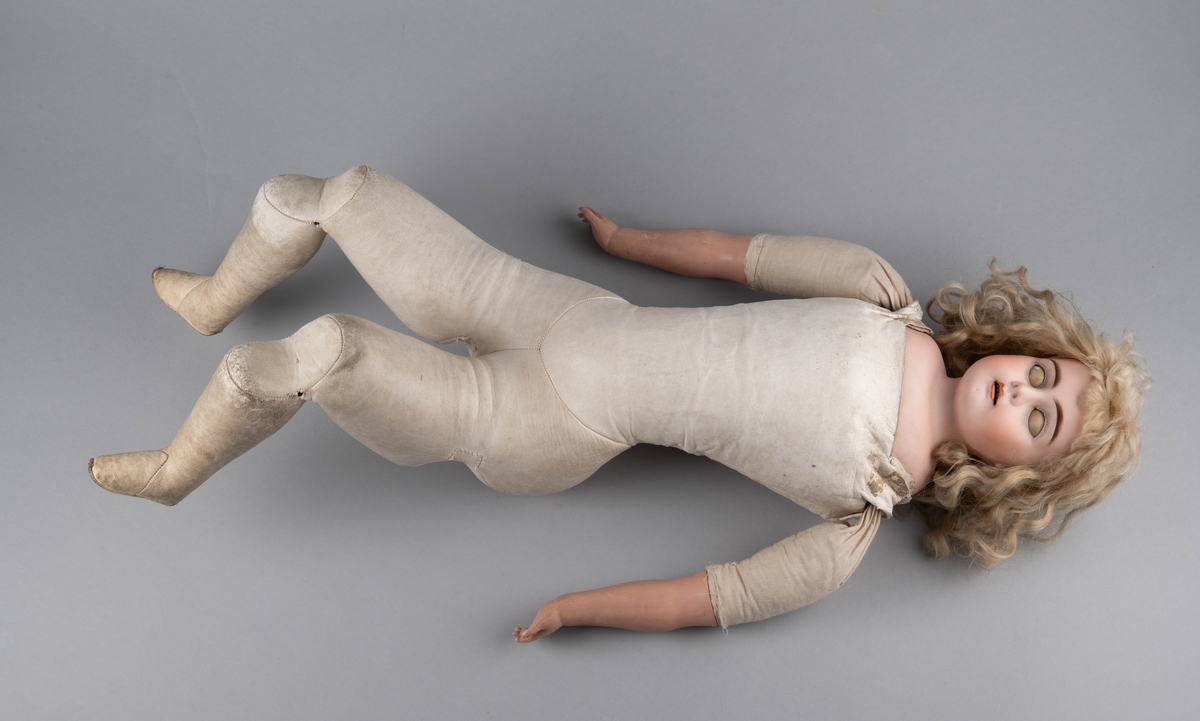 Dukke med porselenshode og underarmer av celluloid. Langt lyst, krøllet hår og blå øyne med bevegelige øyelokk. Stoppet kropp trukket med hanskeskinn, overarmene med tekstil.