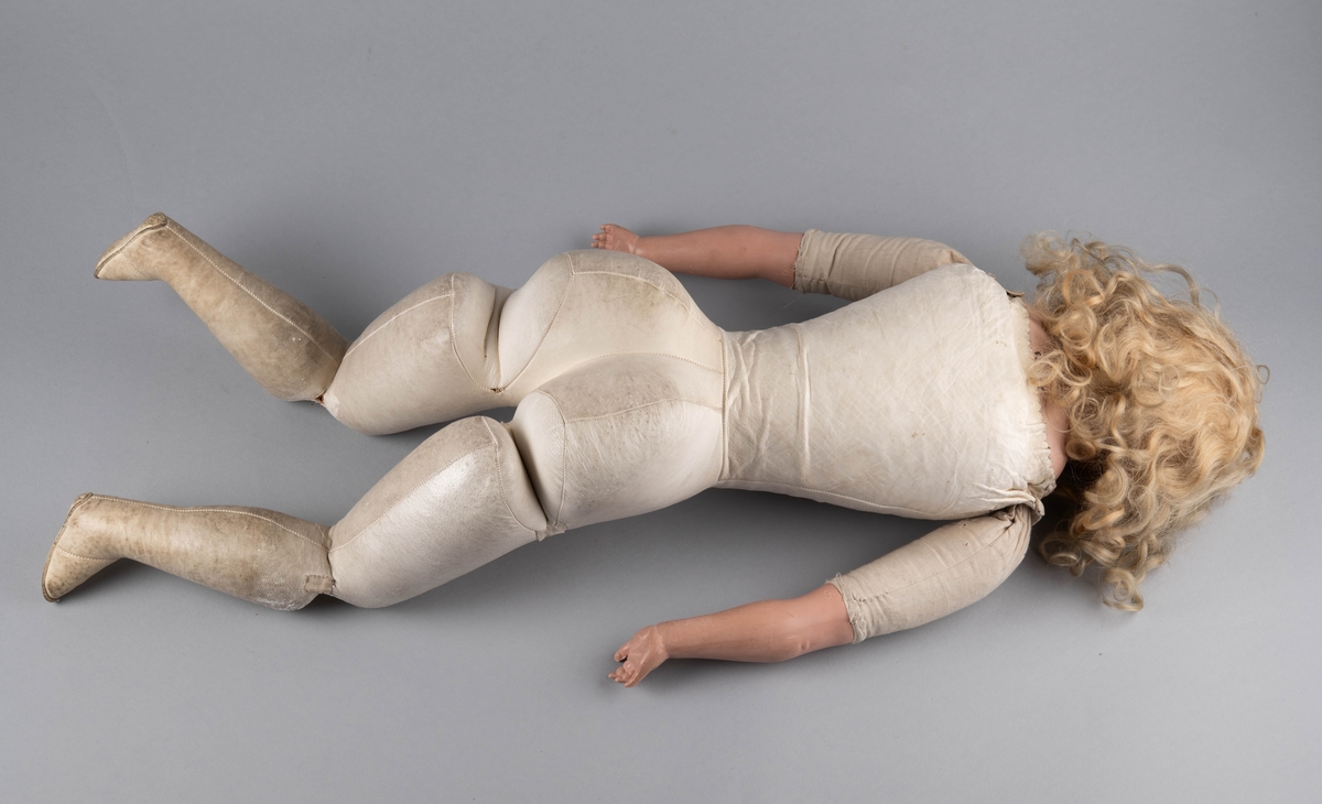 Dukke med porselenshode og underarmer av celluloid. Langt lyst, krøllet hår og blå øyne med bevegelige øyelokk. Stoppet kropp trukket med hanskeskinn, overarmene med tekstil.