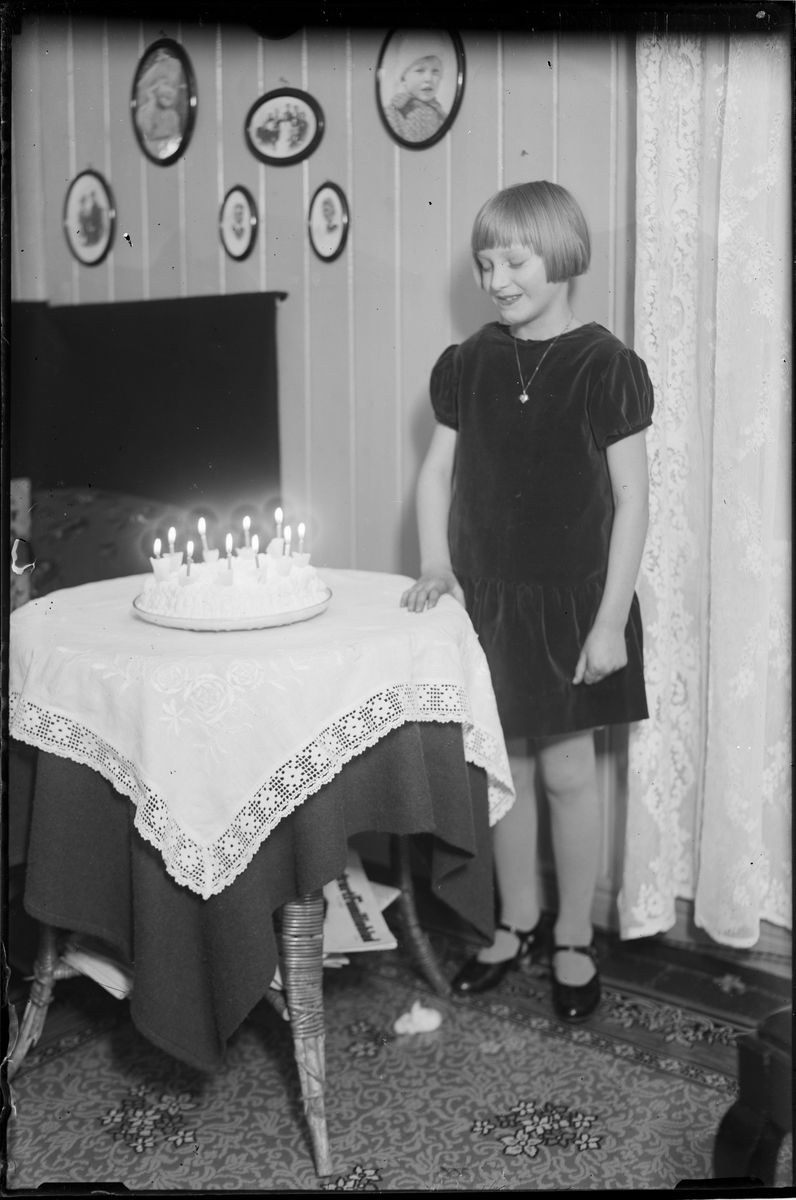 Jente stående ved bord. På bordet står en bløtkake med 10 levende lys. Bilder på veggen i bakgrunnen.