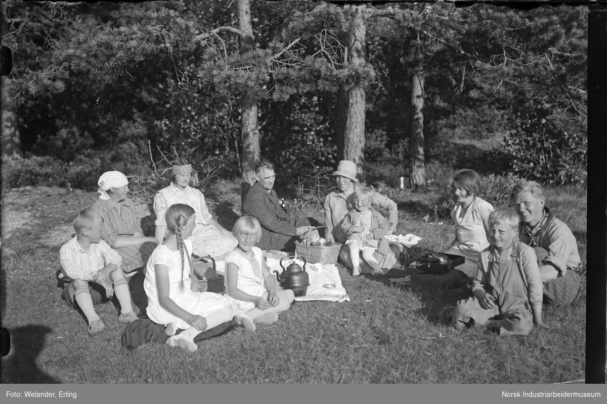 Gruppe mennesker på piknik i naturen. Kurver med mat og drikke er satt ut mellom menneskene og duk på gresset er lagt ut. Kaffekjele står på duken. Platespiller står i gresset mellom menneskene.