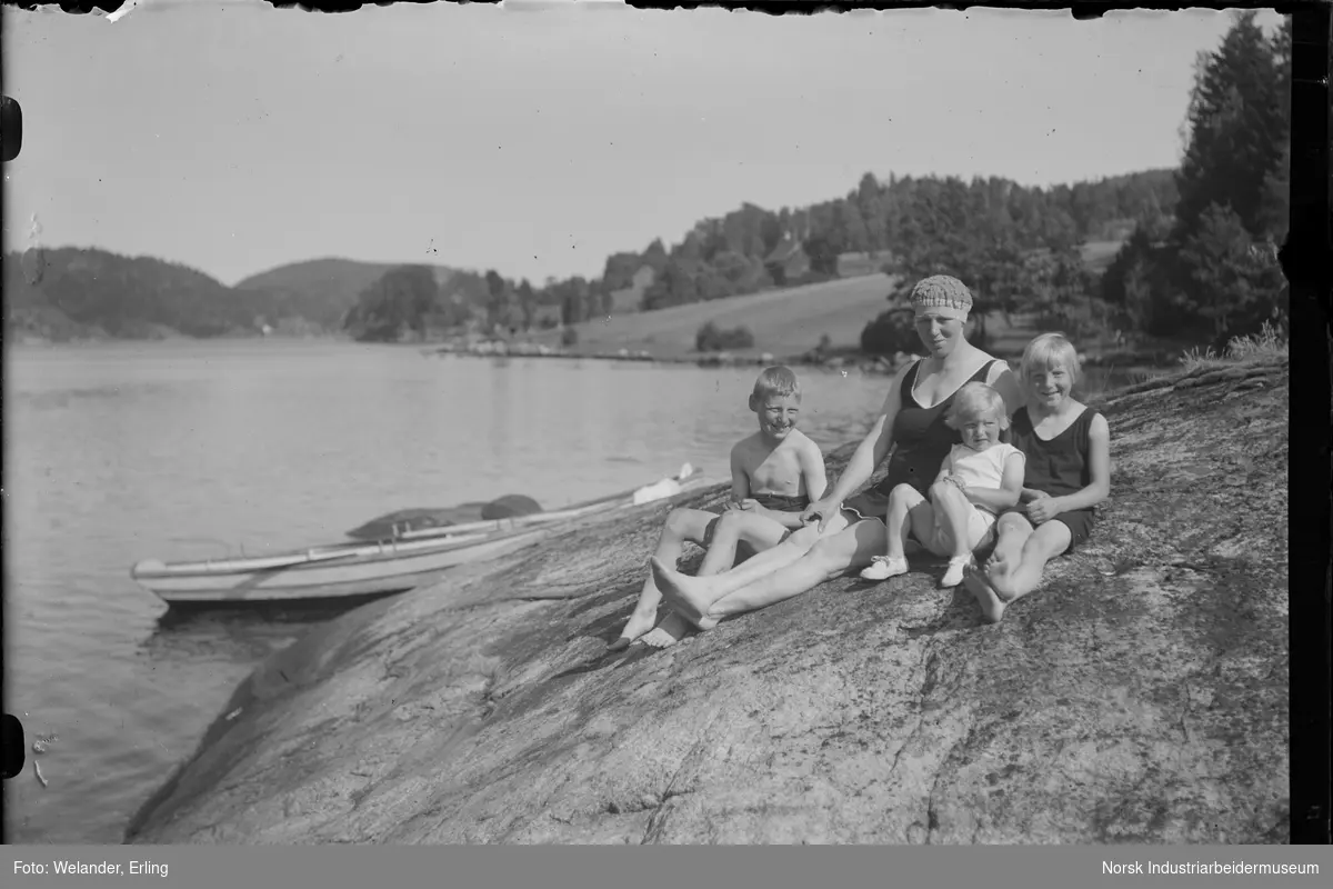 Kvinne og tre barn sittende på svaberg ved vann. Alle kledd i badetøy, kvinnen med badehette på hodet. Robåt liggende i vannkanten. Gårder i bakgrunnen.