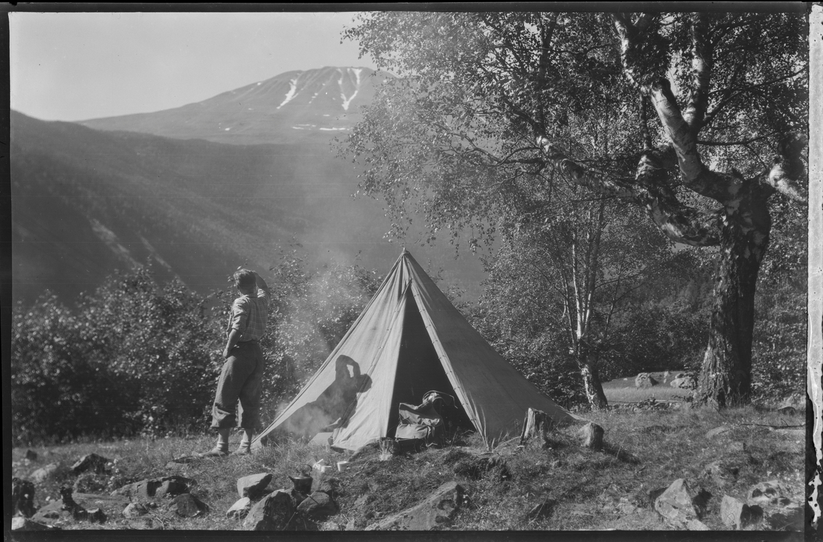 Mann på teltur med utsikt mot Gaustatoppen. Pyramidetelt slått opp i en lysning. Kaffekjelen stående på bål. Sekk stående i teltåpningen.